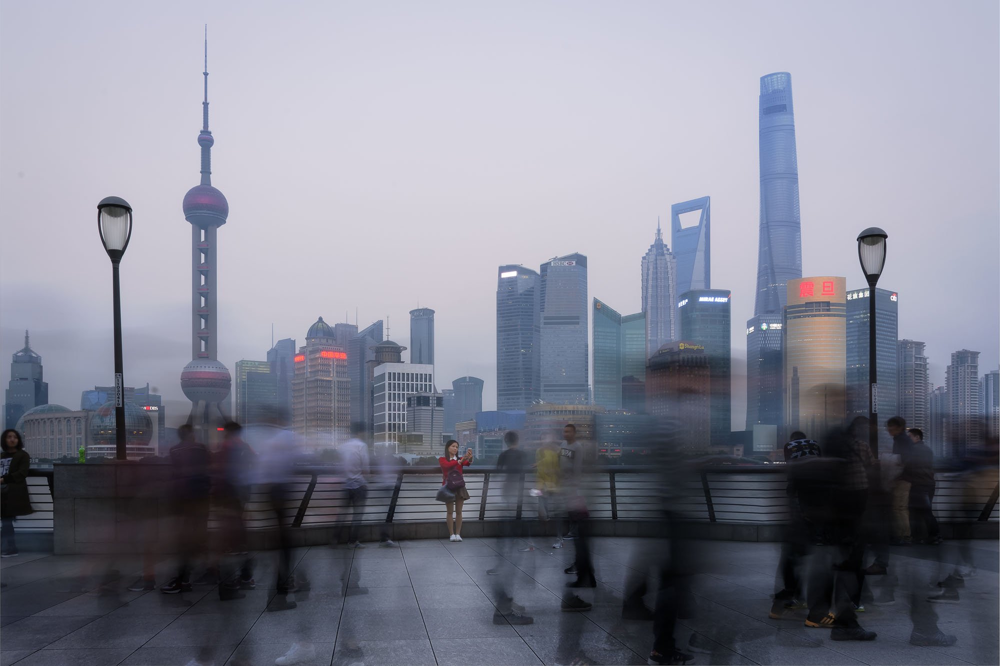 A long-exposure view of the Shanghai skyline across the Huangpu River, with motion-blurred commuters streaming past a single still woman in a red jacket