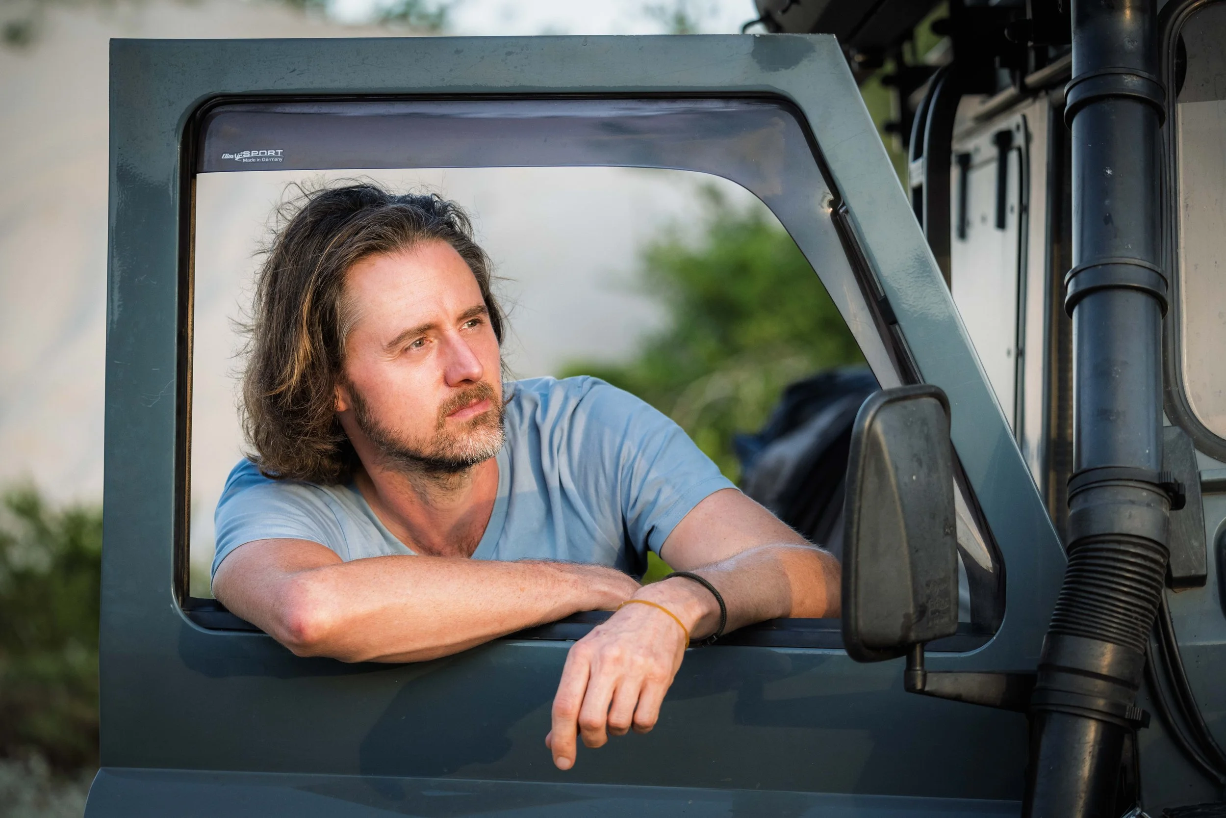 A man with shoulder-length hair and a beard leaning on a vehicle's window frame, looking thoughtfully into the distance.