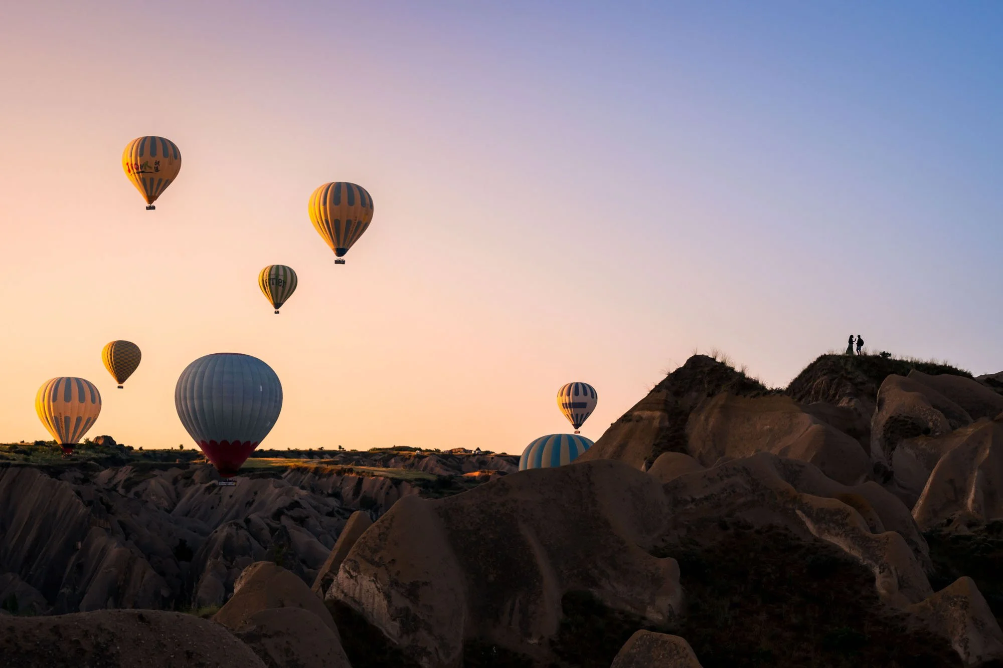 A couple chat on the rocky rim of a deep canyon in Cappadocia, Türkiye, overlooking dozens of colourful hot air balloons rising at dawn.