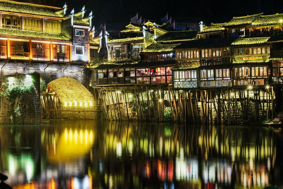 Illuminated traditional wooden stilt houses glowing at night in Fenghuang, China, casting vibrant, liquid reflections across the calm, dark river surface