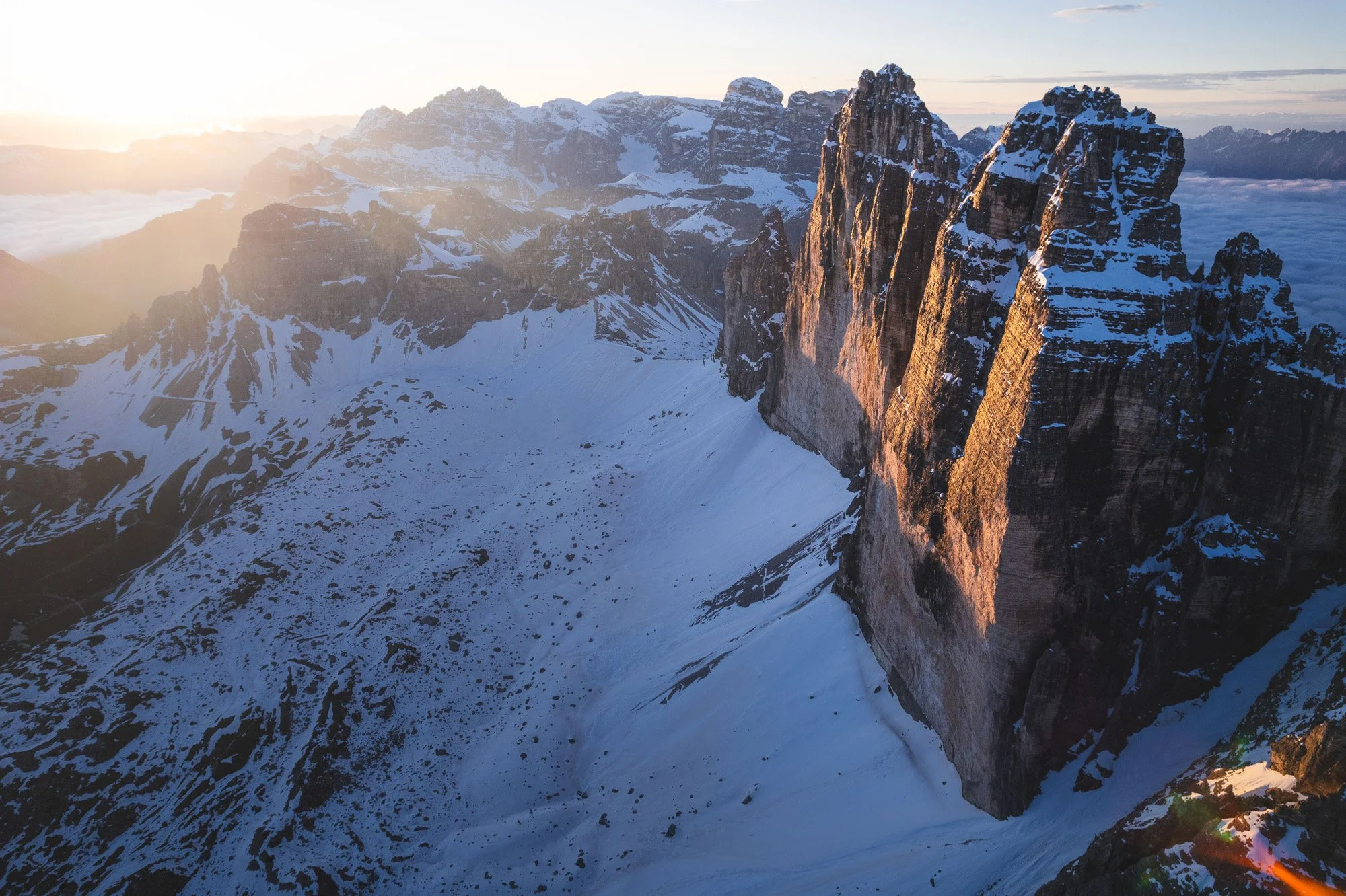 A spectacular aerial drone view of the Tre Cime di Lavaredo in the Dolomites, Italy, at sunrise. The immense scale and dramatic side-lighting trigger a psychological feeling of awe and environmental exploration.