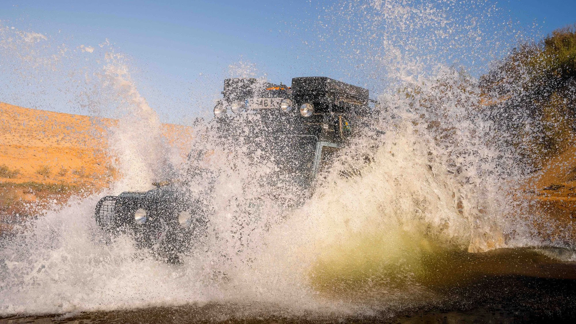 A Land Rover Defender aggressively crossing a desert riverbed, creating a massive wave of white water. The explosive splash represents the adrenaline of the search and the physical cost of the moment.