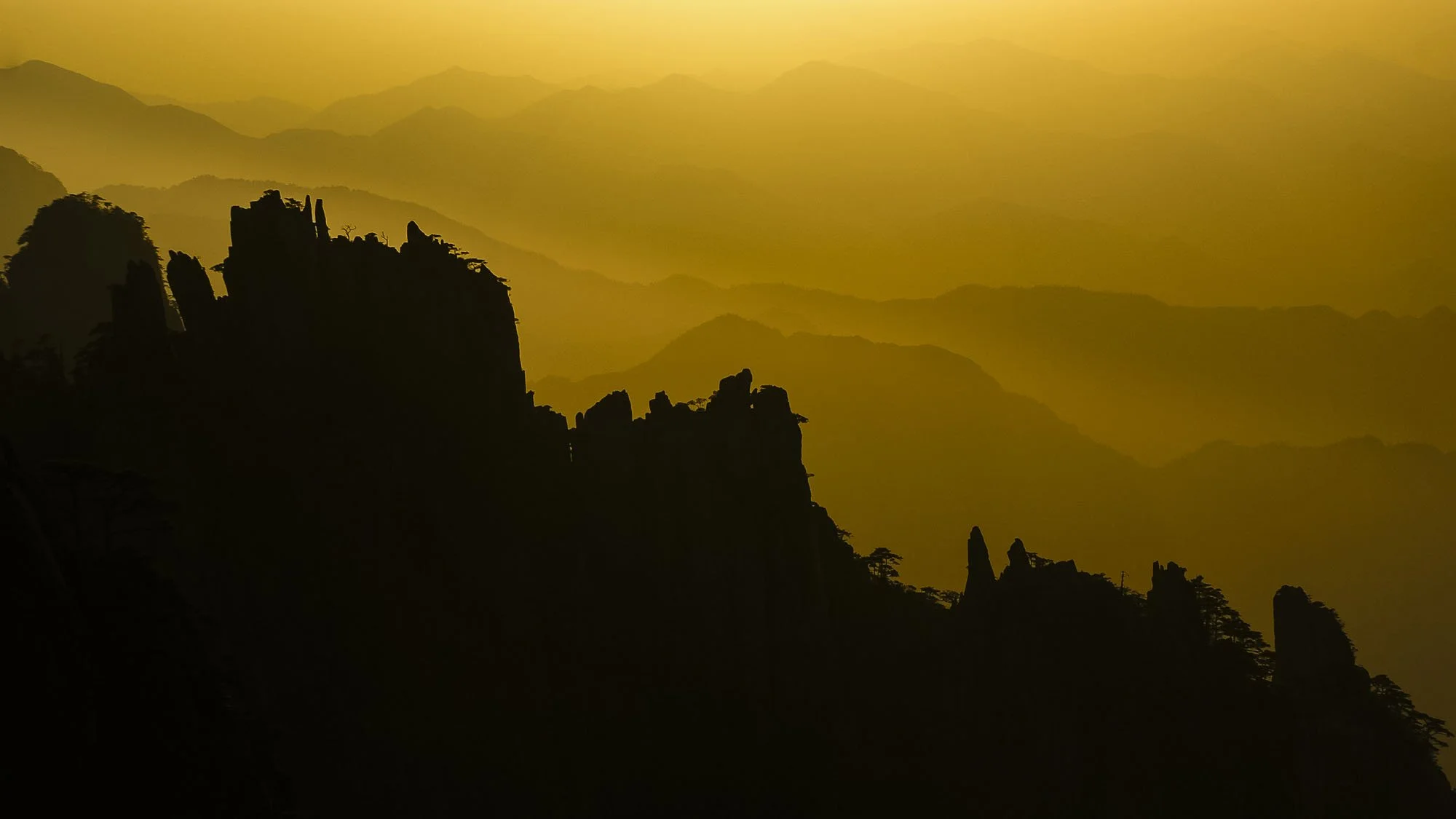 Jagged black mountain peaks in pure silhouette rising through golden morning mist and atmospheric haze at sunrise in China.