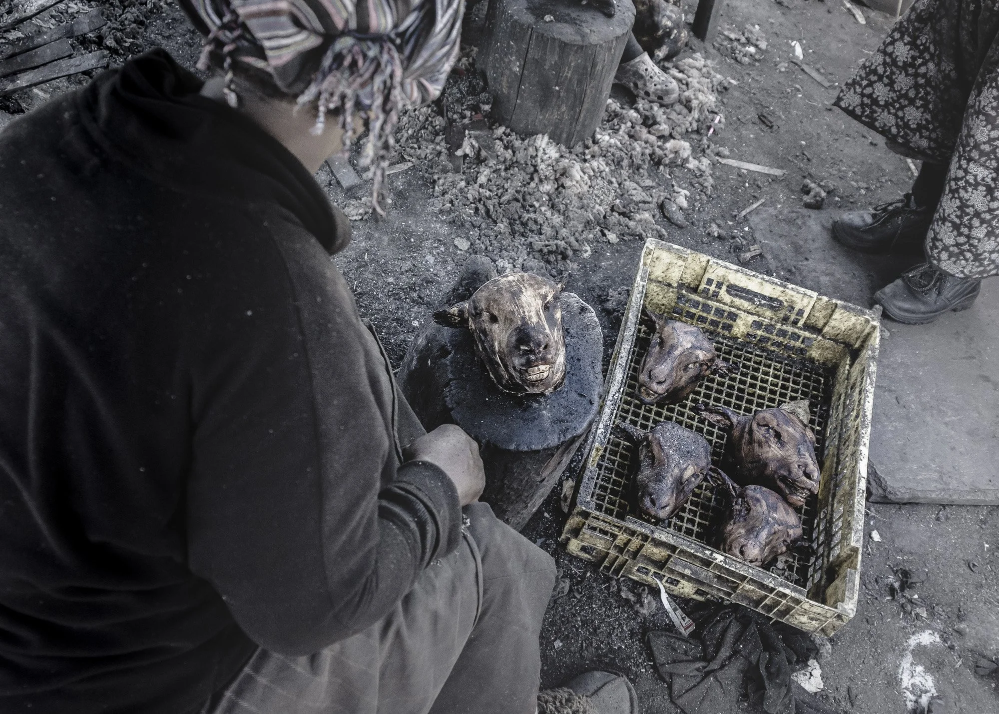 A person is sitting next to a makeshift blacksmithing station with a scaffold, welding or forging animal heads, likely sheep or goat, which are placed in a plastic crate and on a blackened surface. The setting appears gritty and industrial.