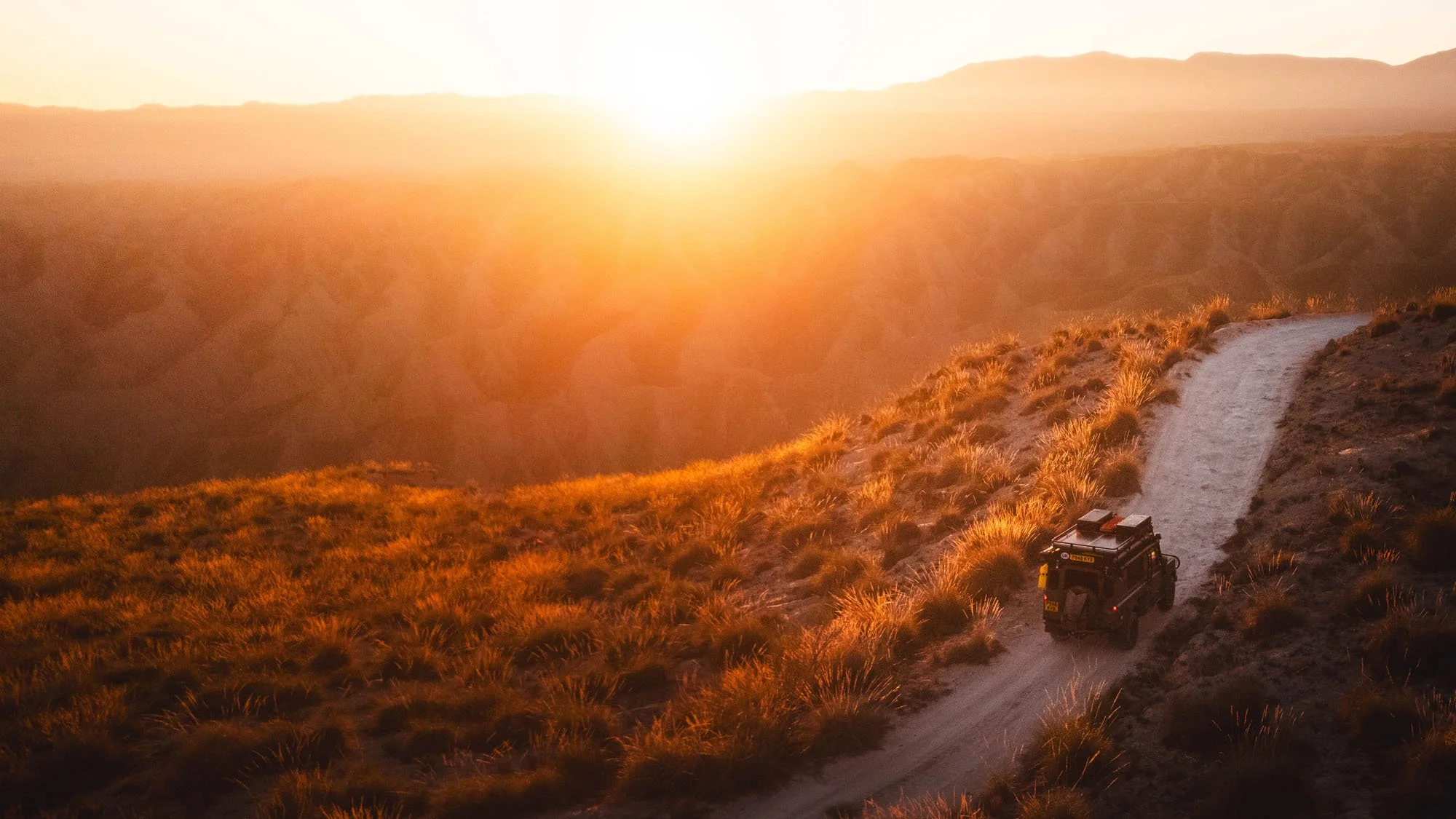 A Land Rover Defender driving along a winding mountain ridge track overlooking a vast, misty valley glowing in golden sunset light