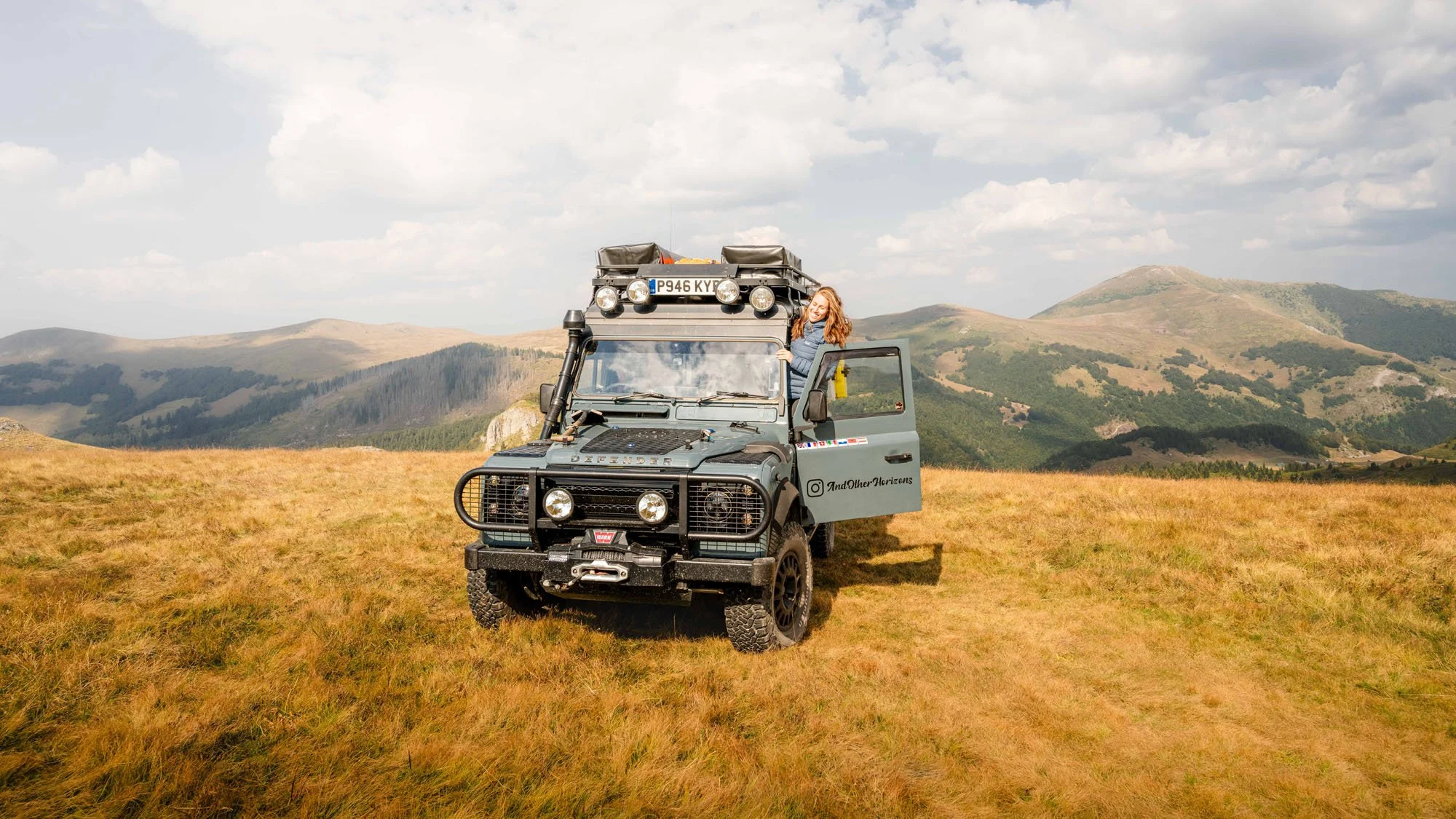 A woman smiles from the side step of an expedition Land Rover Defender in the mountains. The presence of the vehicle and the subject's expression provide 'Visual Breadcrumbs' for the viewer to construct their own story