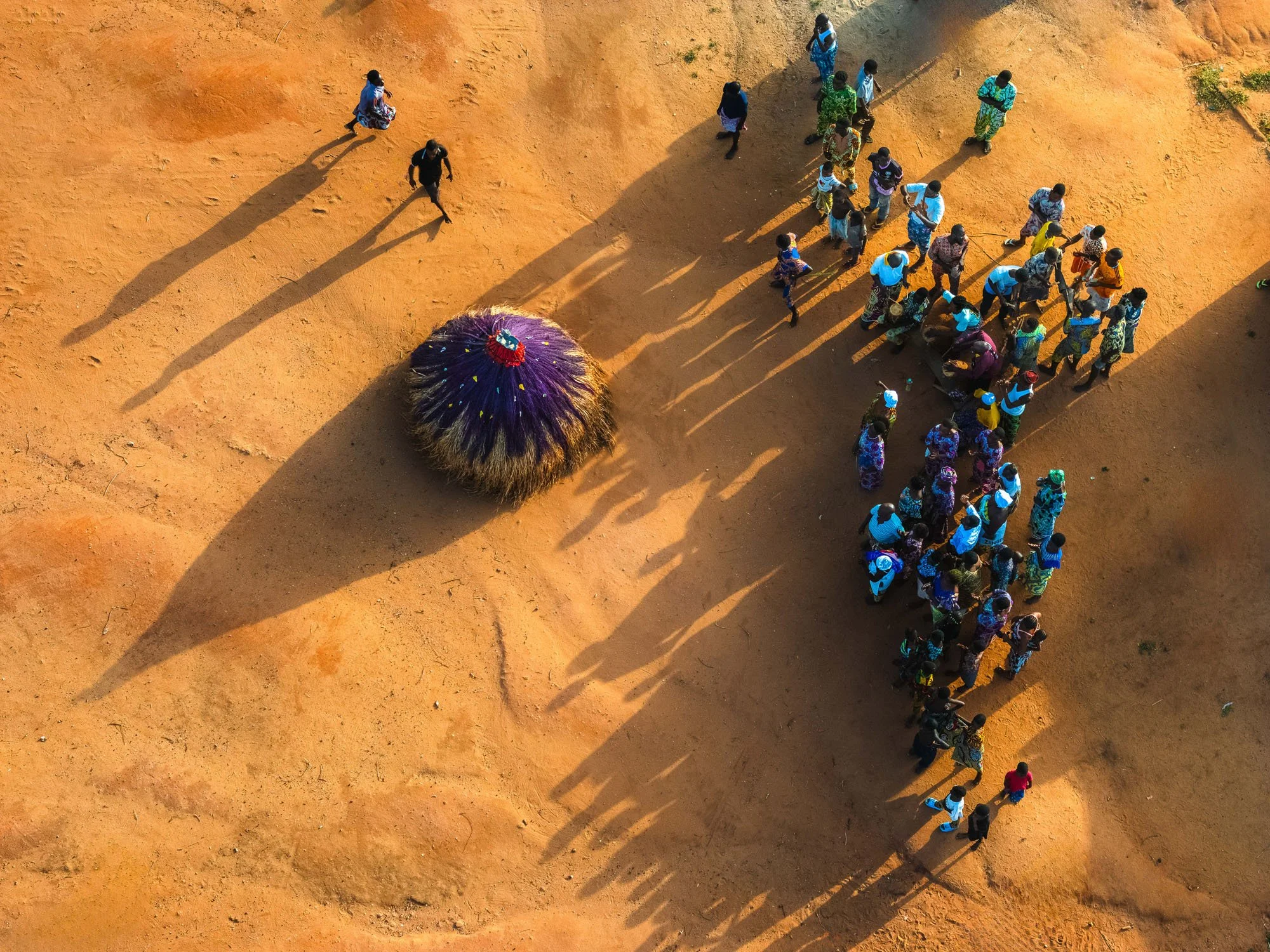An aerial drone view of a conical straw Zangbeto casting a long shadow across red dirt, facing a gathering of villagers on the shores of Lake Ahémé, Benin