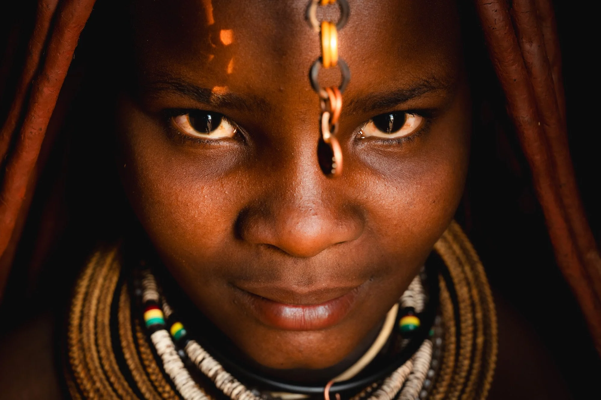 Close-up portrait of an African woman wearing traditional beaded necklaces and a metallic forehead ornament, with a focused expression.