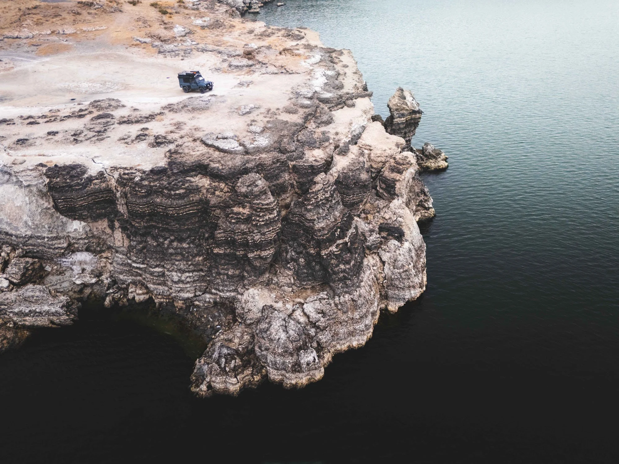 Aerial drone view of a Land Rover Defender parked on a rugged, eroded cliff edge above deep water.