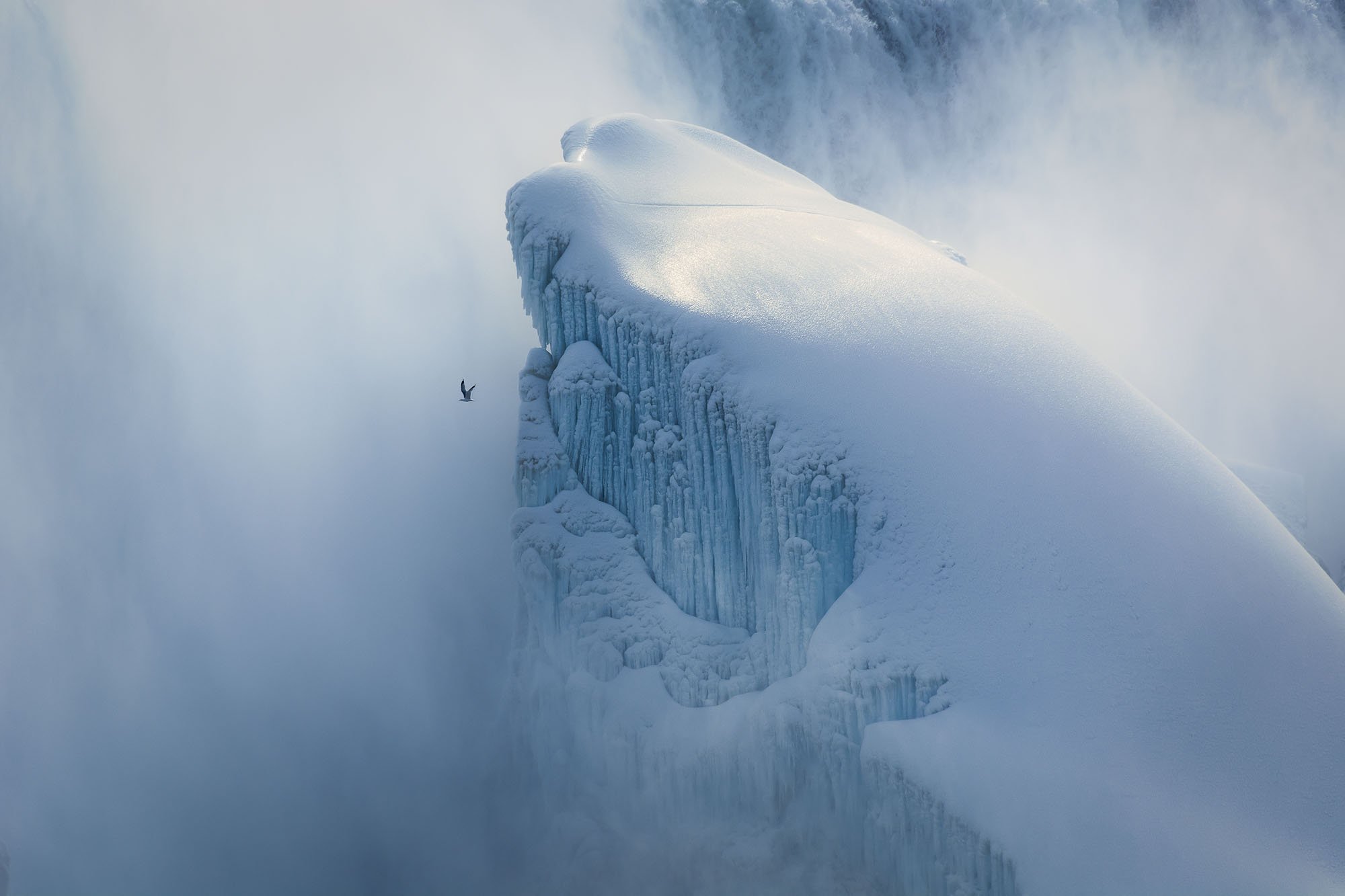 A colossal, frozen blue-and-white ice monolith shrouded in winter mist at Niagara Falls, with a solitary bird flying past to emphasize the massive natural scale