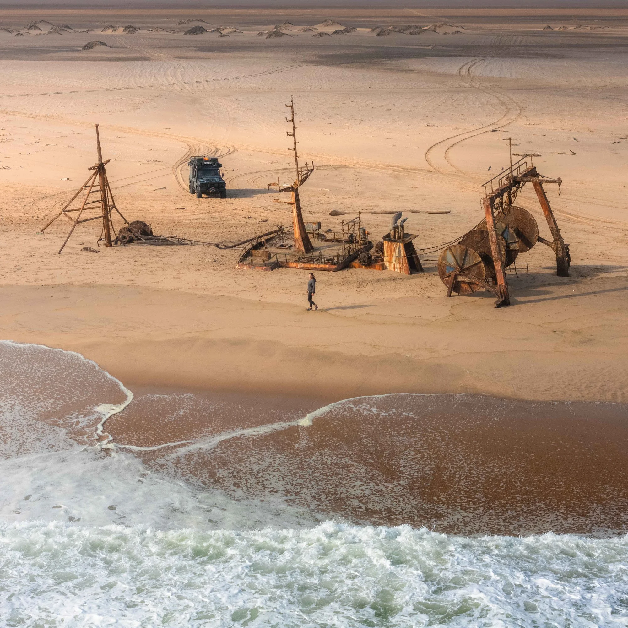 A beach scene with abandoned rusted oil rigs and a vehicle, with a person walking on the sand near the water.