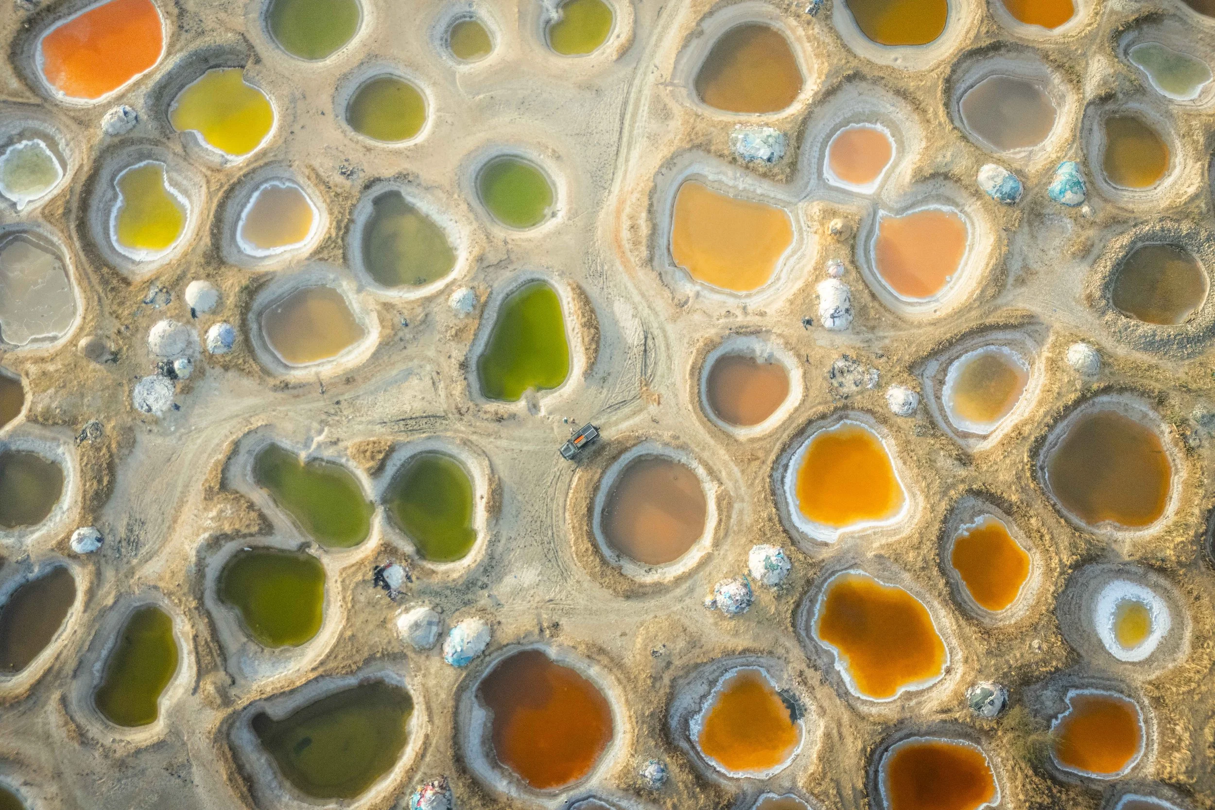 An aerial view of multiple colorful salt evaporation ponds with vibrant green, yellow, orange, and brown hues, separated by narrow dirt pathways.