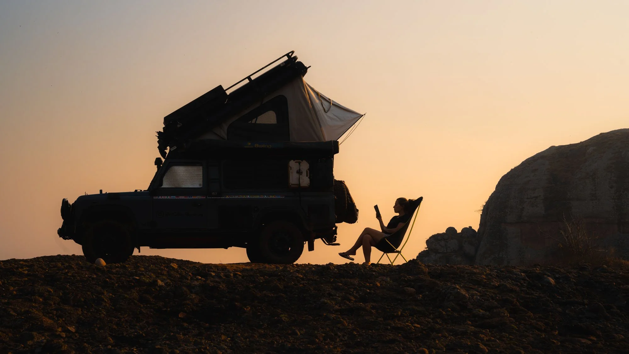 A silhouette of an expedition Land Rover Defender with a roof tent and a traveler reading in a camp chair on a ridge at sunset in Angola.