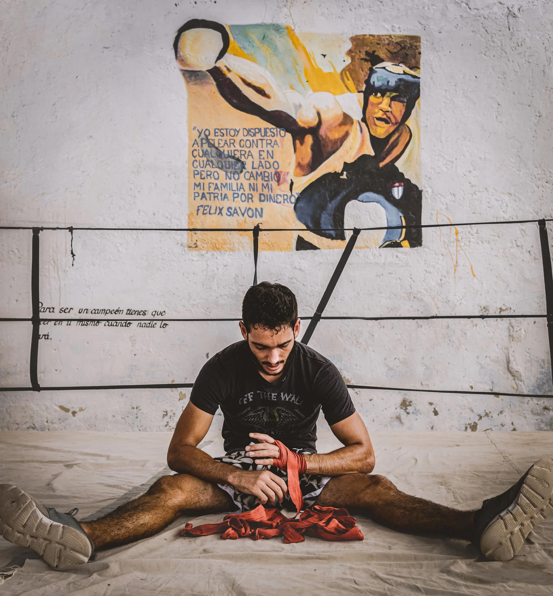A young man sitting on the floor of a boxing gym, tying his red hand wraps. Behind him, a mural of a boxer and motivational quote on the wall.