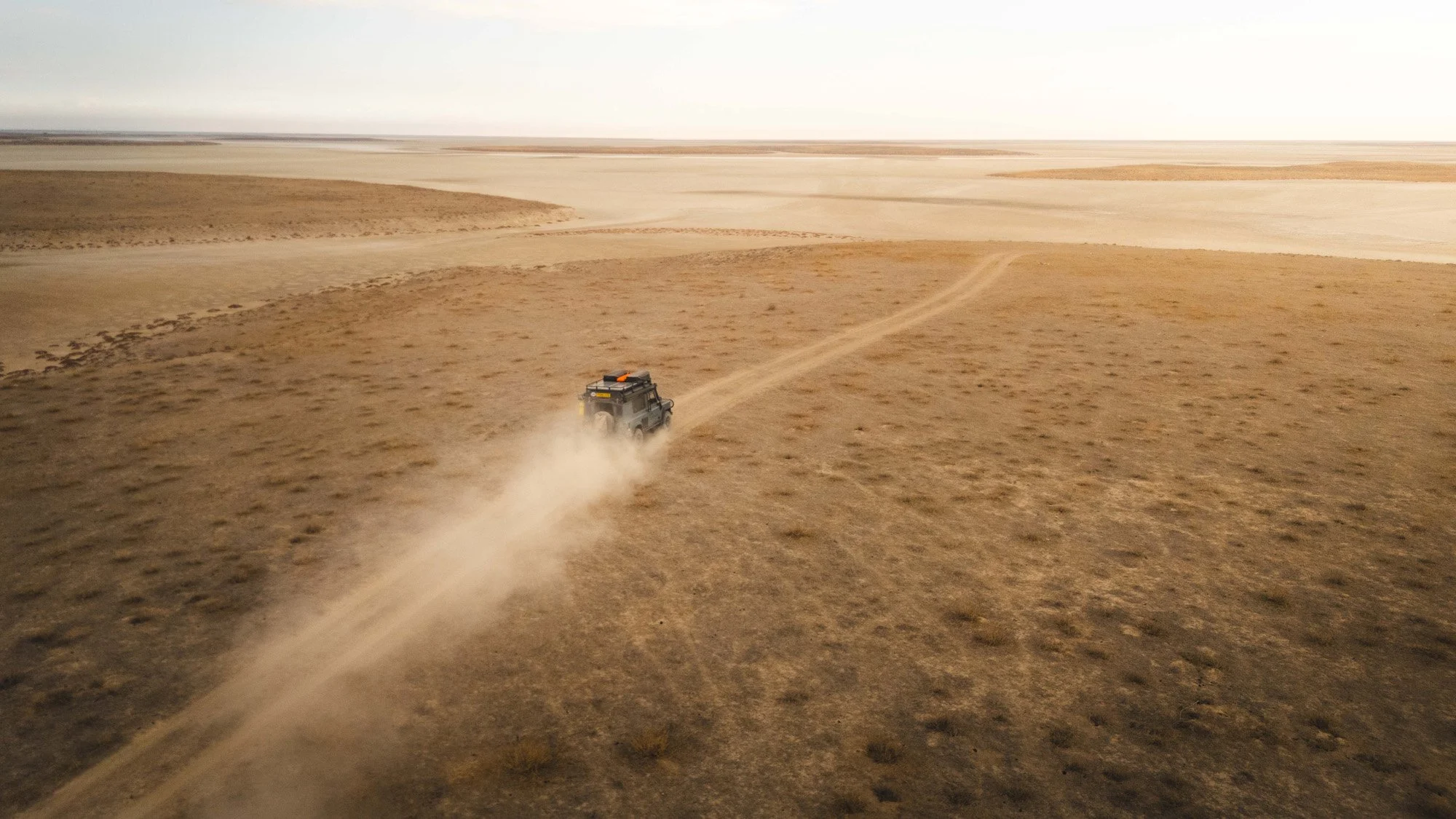 An aerial drone shot of a Land Rover Defender kicking up a long trail of dust as it drives across the vast, empty salt plains of Türkiye. The trail acts as a powerful leading line, emphasizing the scale of the journey.