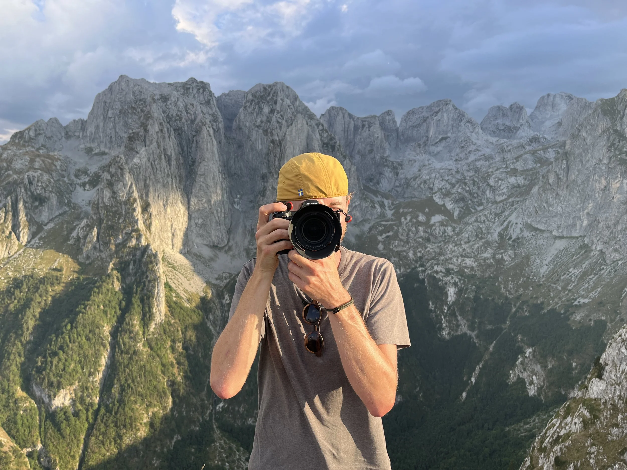 Person taking a photo with a camera in front of a mountain landscape with rocky peaks and green valleys.