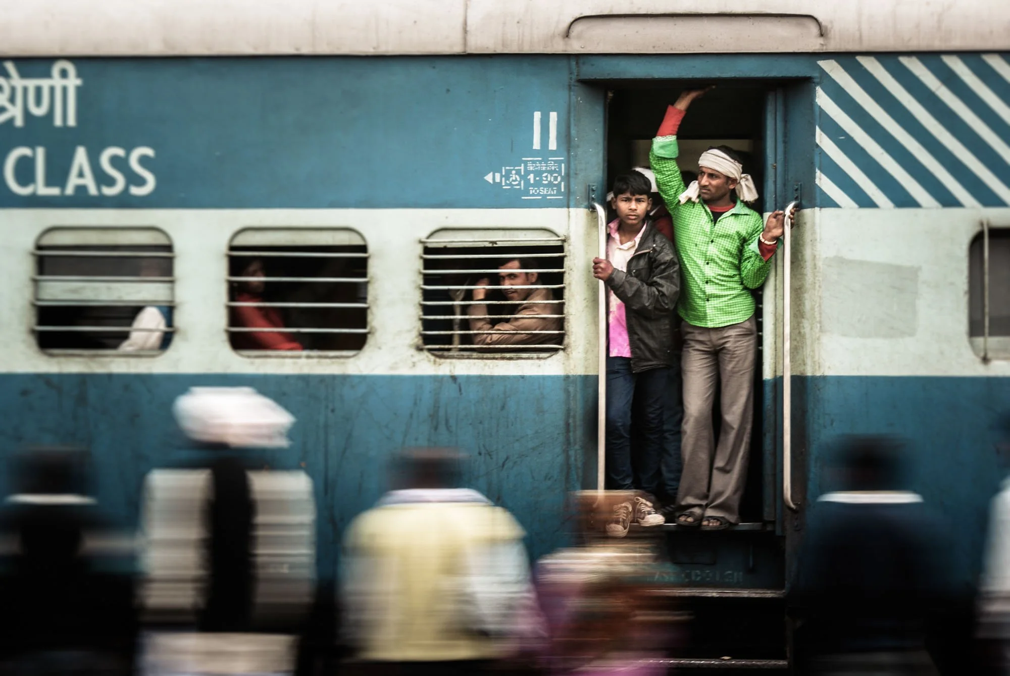 Passengers framed in the open blue doorway of an Indian railway train carriage in Rajasthan, surrounded by a motion-blurred crowd on the platform.