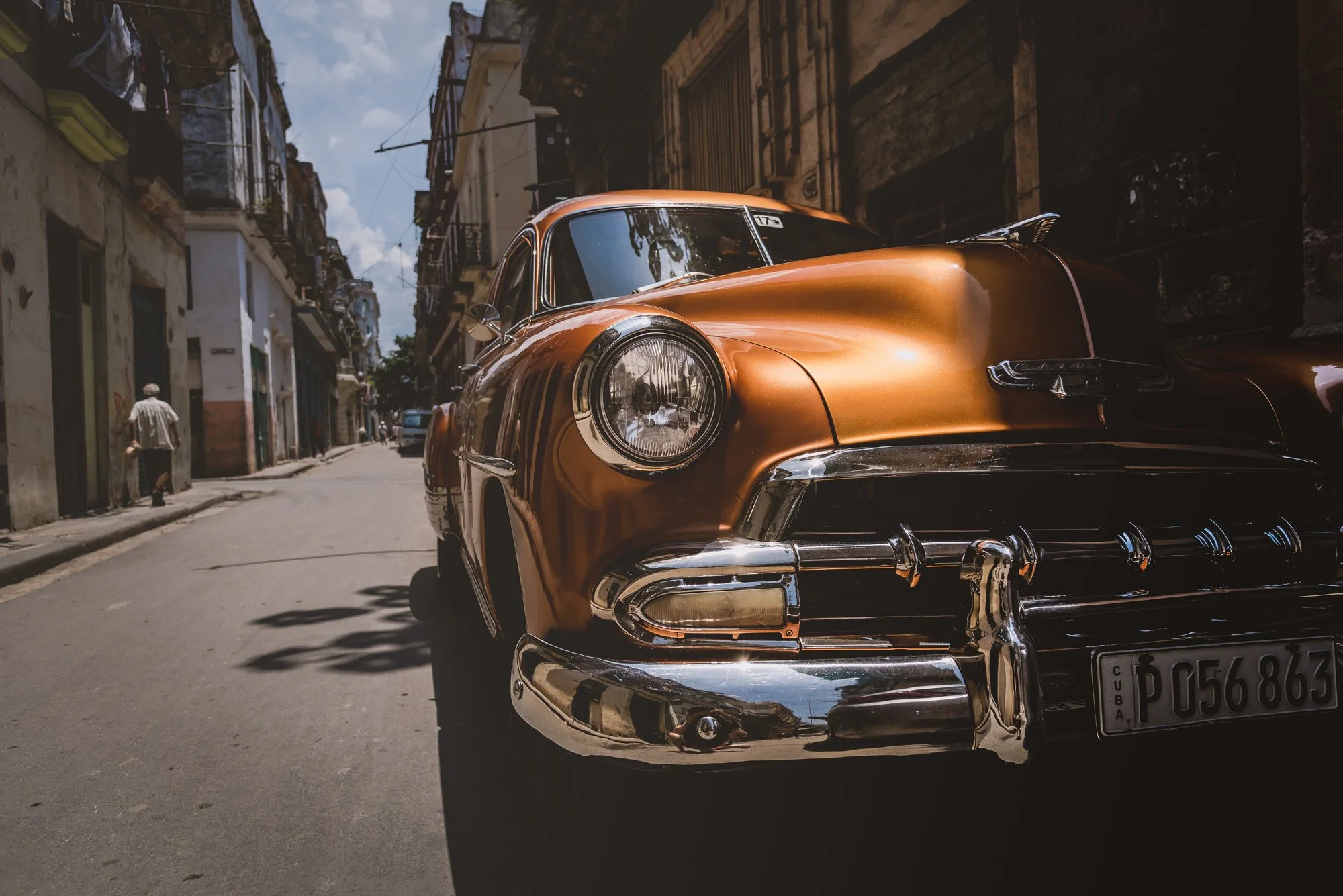 A vintage copper-colored car parked on a narrow street in Cuba, with old buildings and a person walking by in the background.