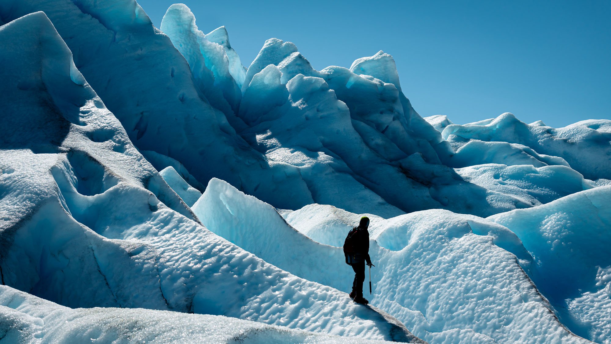 A lone hiker silhouetted against the colossal, deep blue ice seracs and jagged crevasses of the Perito Moreno Glacier in Patagonia, Argentina