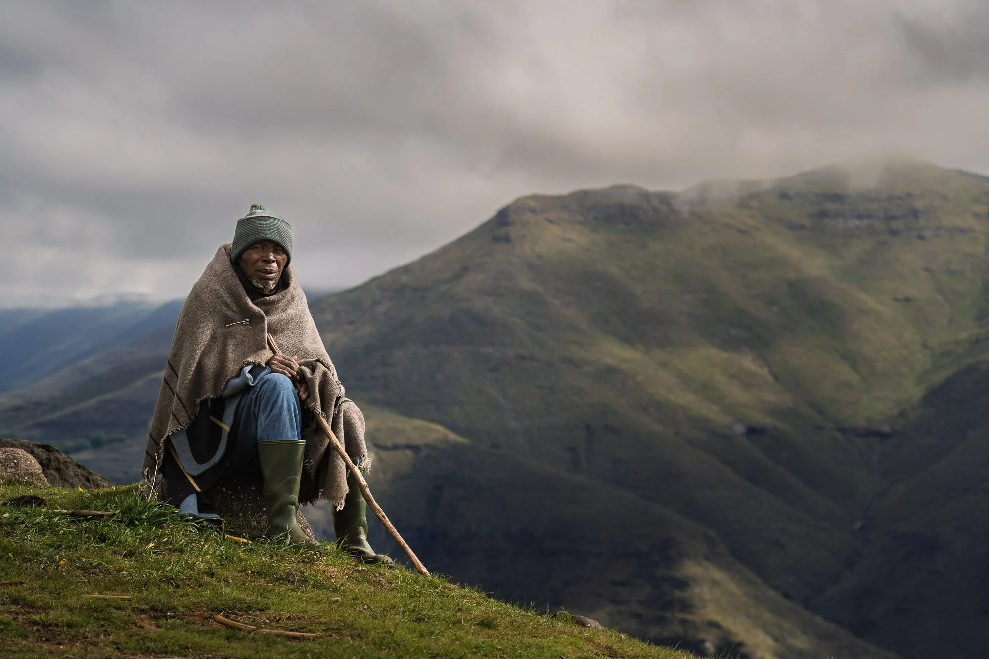 A wide-angle portrait of a Basotho farmer in a traditional wool blanket and rubber boots sitting on a hillside in Lesotho, demonstrating how geography and friction dictate visual composition.