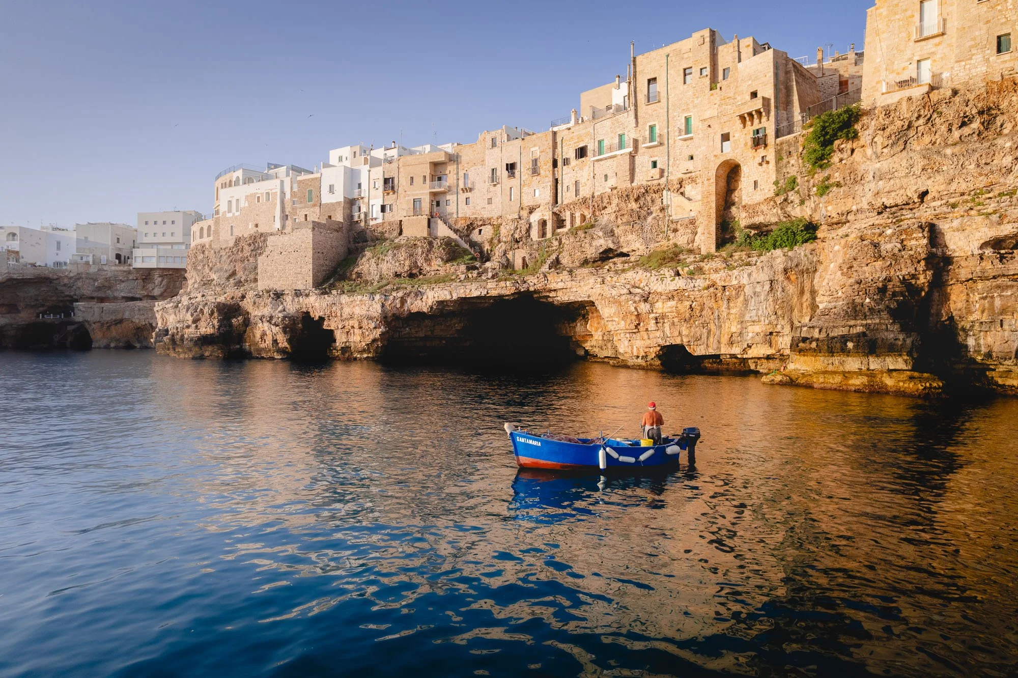 A lone fisherman in a blue boat beneath the massive limestone cliffs and white architecture of Polignano a Mare, Italy, demonstrating how the dark sea cave creates a visual information gap in photography