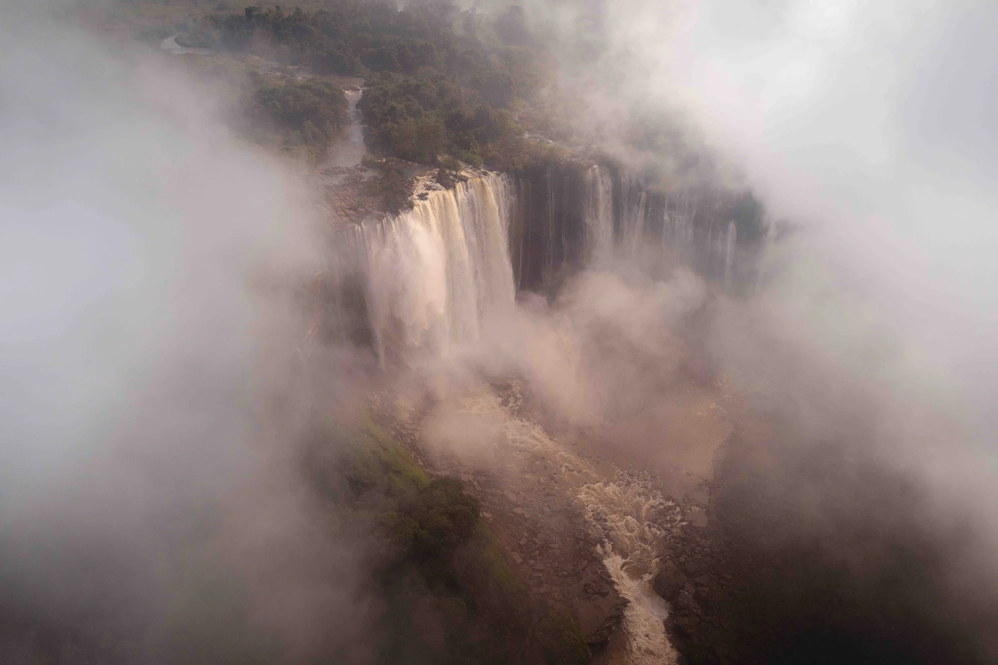 An aerial drone view of the Calandula Falls in Angola partially obscured by mist. The massive scale and implied roar of the water convey overwhelming kinetic energy, commanding immediate, primal attention through the sheer force of nature.