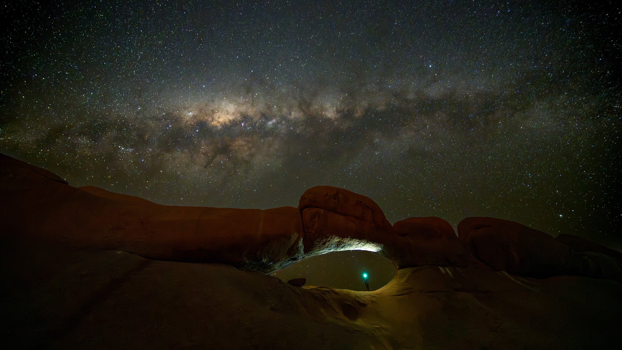 The Milky Way galaxy stretching over a natural rock arch in Spitzkoppe, Namibia. A tiny human figure with a flashlight provides a powerful sense of scale, triggering a psychological response of awe and perspective.
