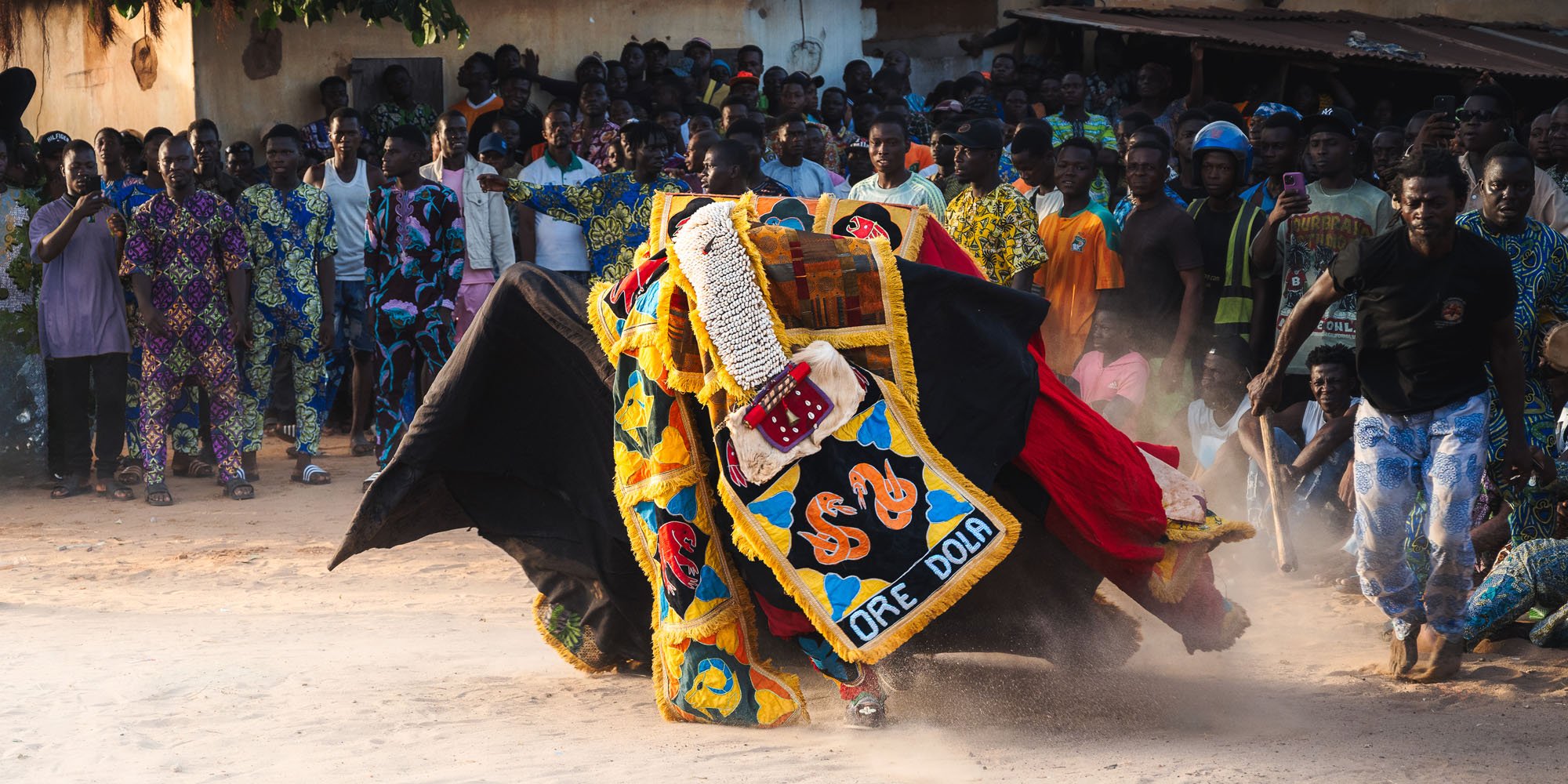 An elaborate Egungun masquerade figure spinning in a crowd in Benin. The 'Ore Dola' panel and the surrounding chaos act as a 'Visual Breadcrumb,' leaving unanswered questions that drive deeper viewer engagement.