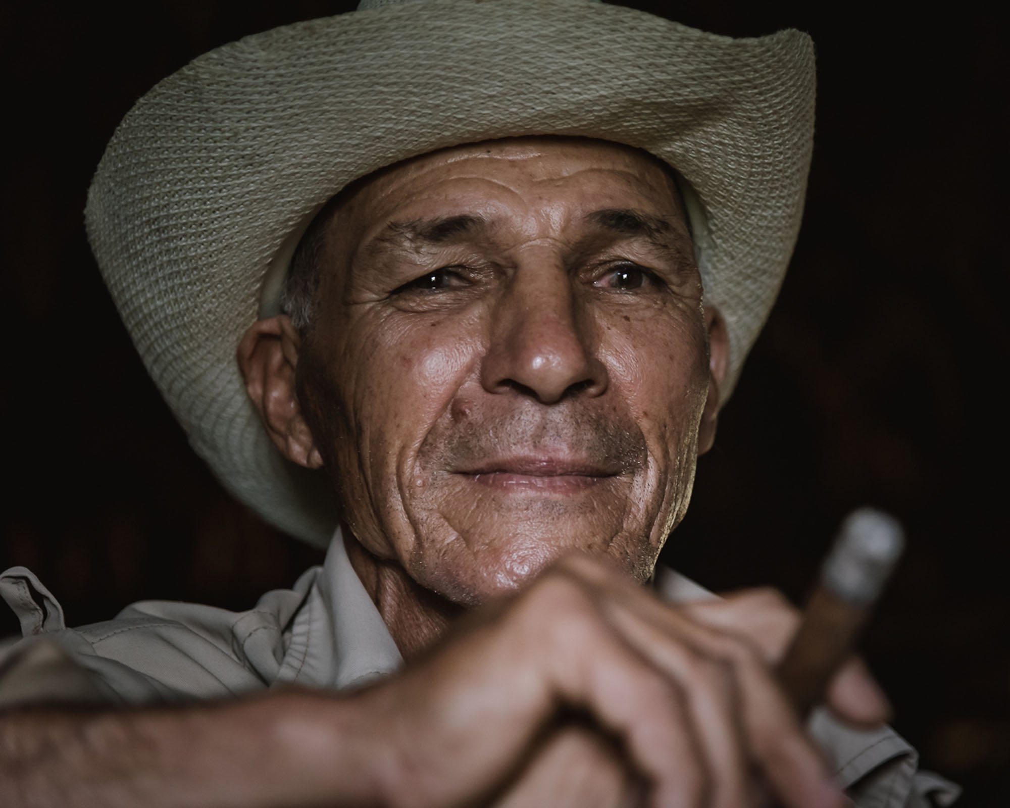 Close-up environmental portrait of an elderly Cuban tobacco farmer in a traditional straw hat holding a cigar under directional barn light in Viñales, Cuba