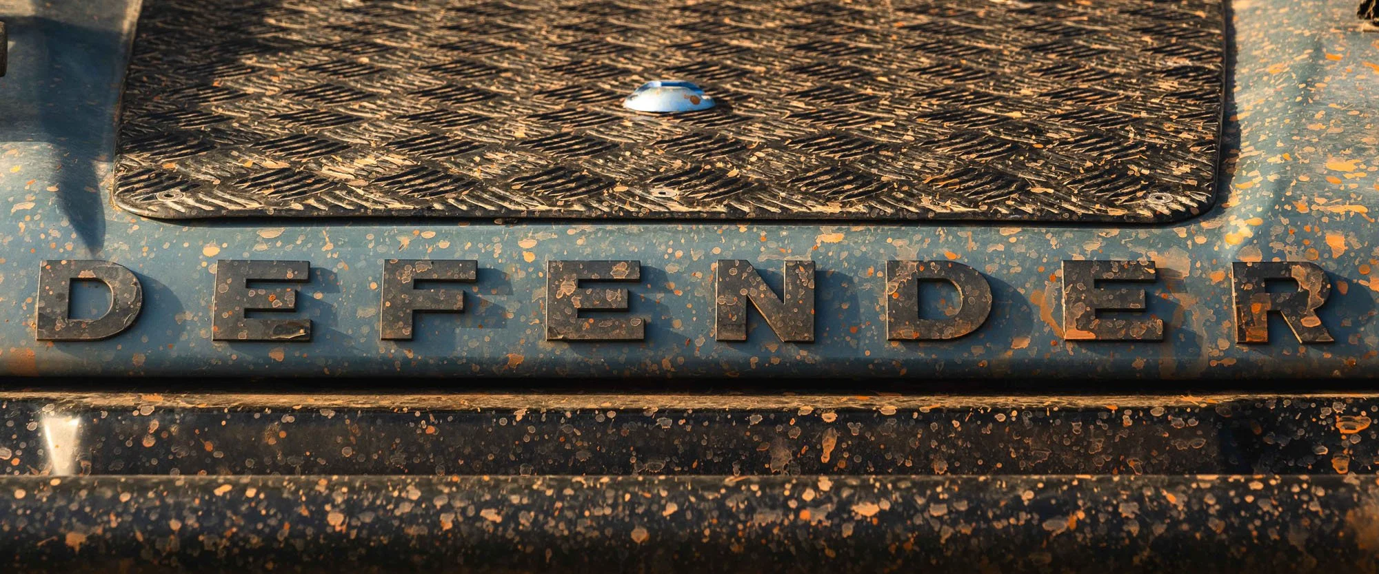 A gritty, close-up study of the 'DEFENDER' badge on a Land Rover covered in fresh orange mud. This serves as a 'biological receipt'—proof of the friction premium and effort required in expedition photography.
