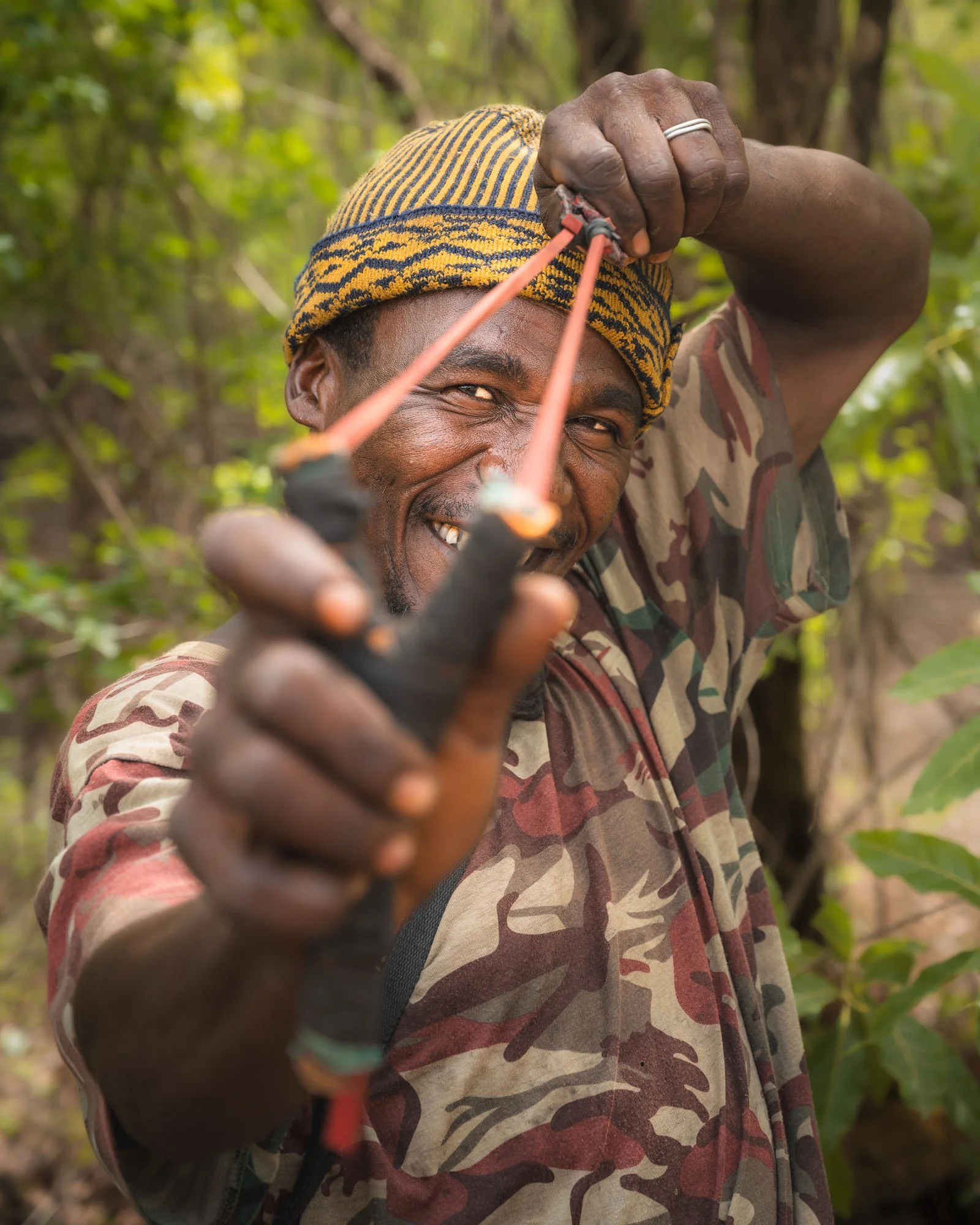 Meaningful photography study: A smiling man in Guinea aims a traditional wooden slingshot directly at the camera lens. illustrates how a non-threatening, empathetic approach creates impactful images