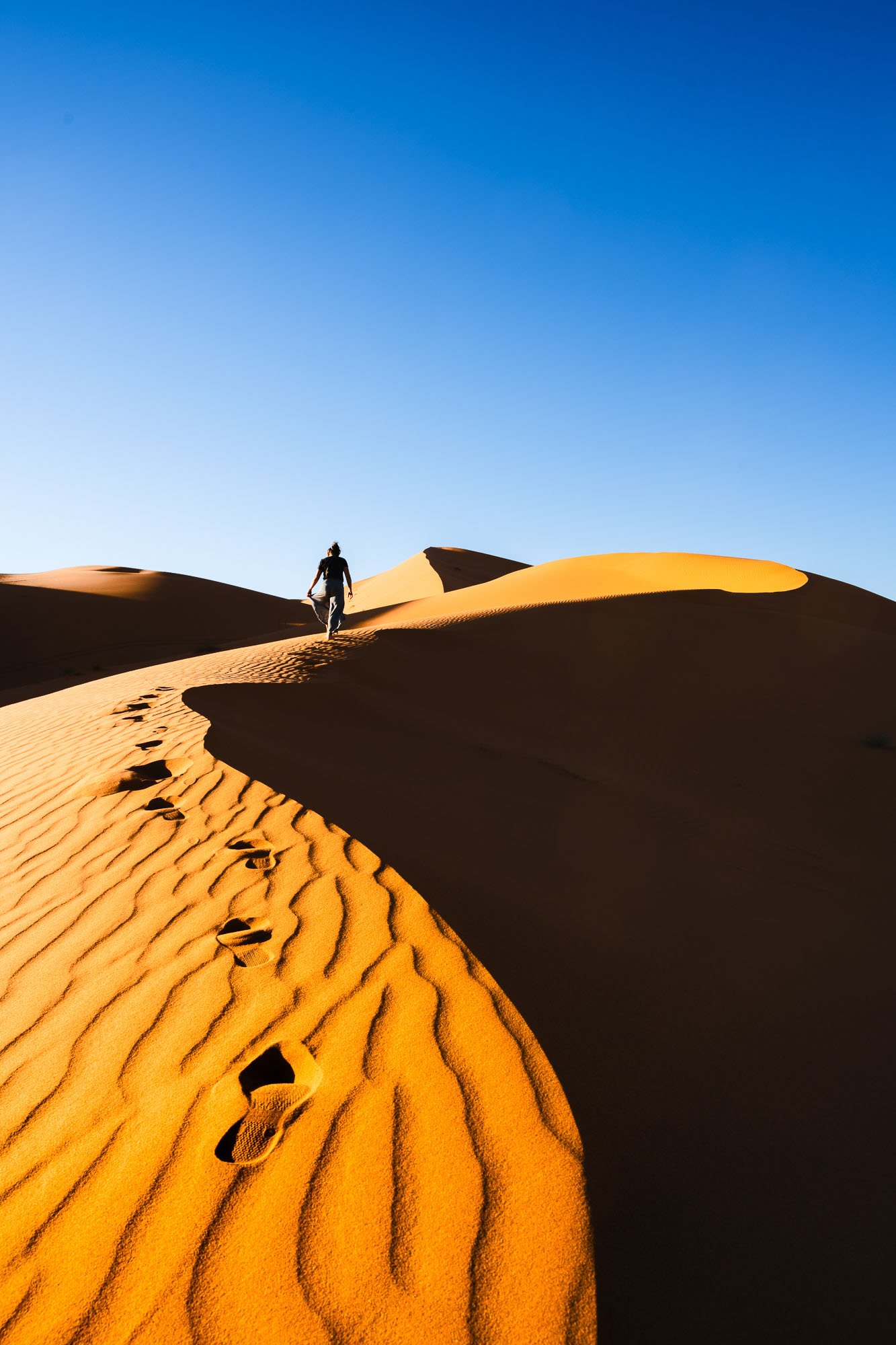 A line of footprints leading up a massive orange sand dune in Merzouga, Morocco, serving as visual breadcrumbs that prove a presence while withholding the destination