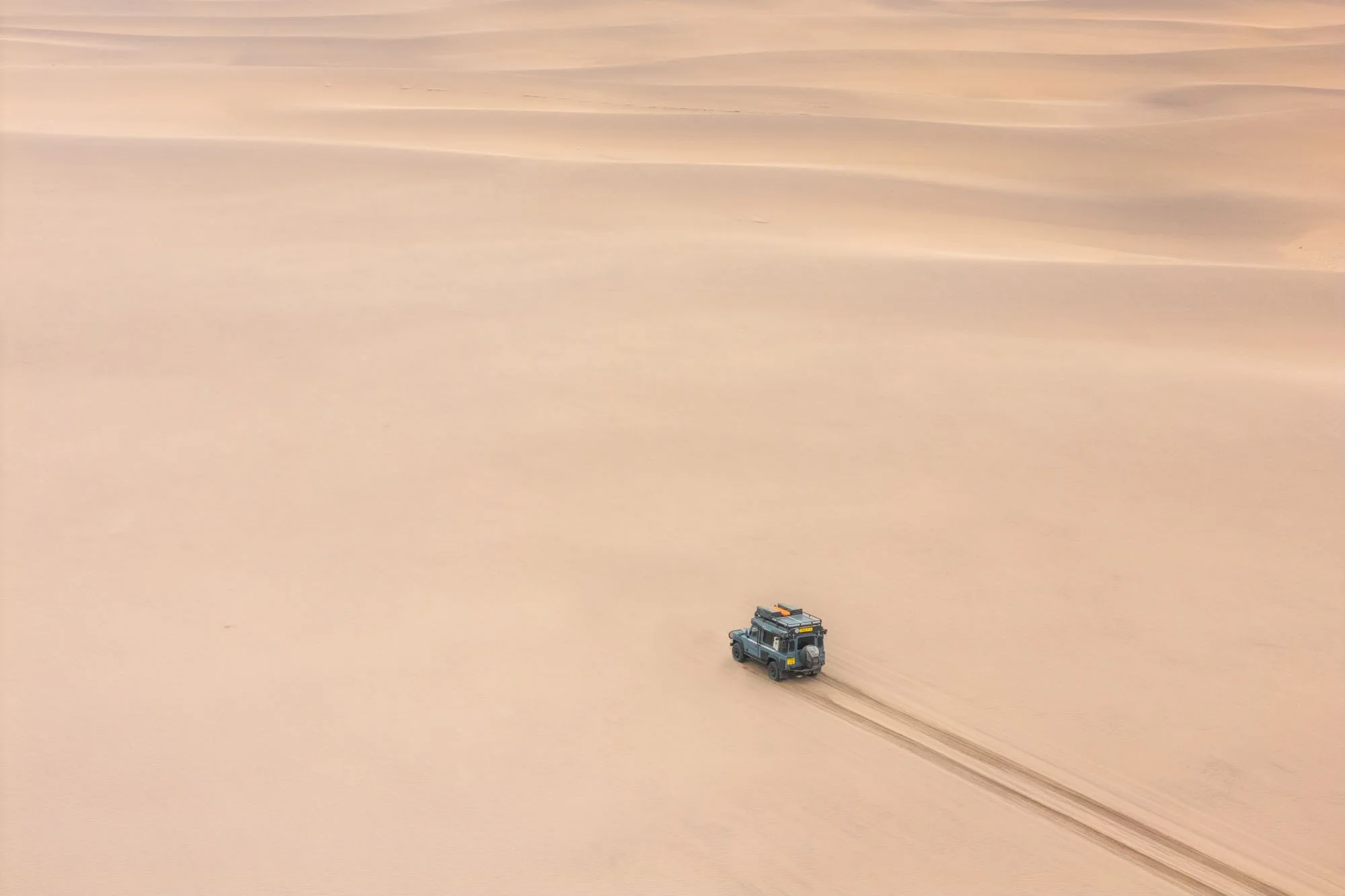 An extreme wide aerial shot of a Land Rover Defender crossing the massive dunes of the Namib Desert. This represents the final directive: stop being a tourist and start being a witness to the world.