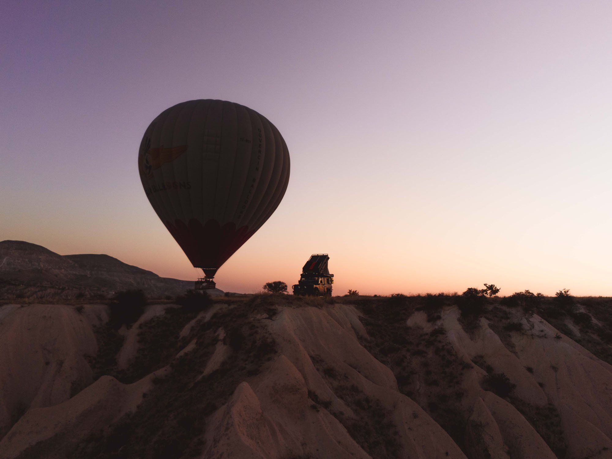 A silhouette of a Land Rover Defender on a ridge in Cappadocia, Türkiye, dwarfed by a massive hot air balloon at dawn, illustrating relational processing in photography