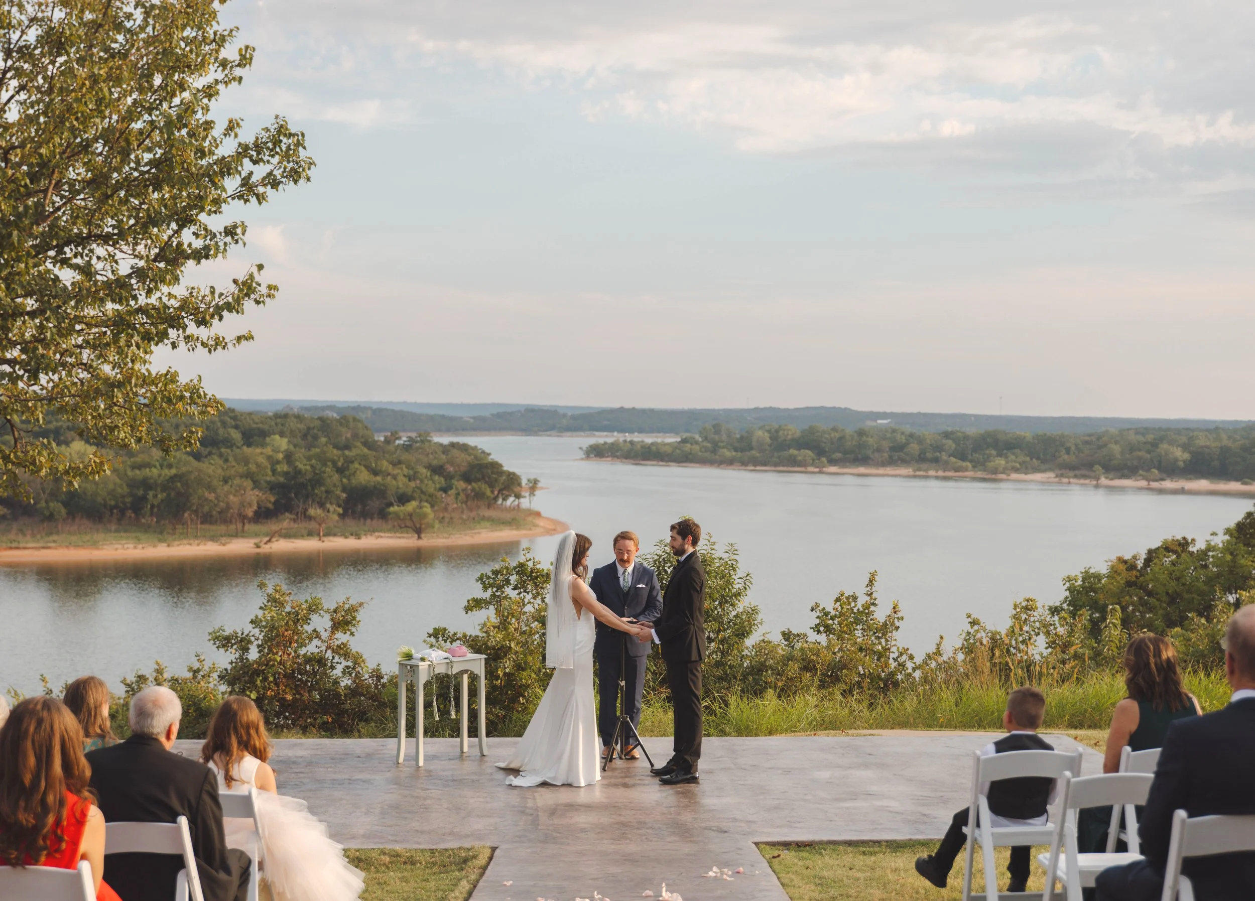 A couple gets married outdoors near a water body, with an officiant, surrounded by seated guests, during daytime.