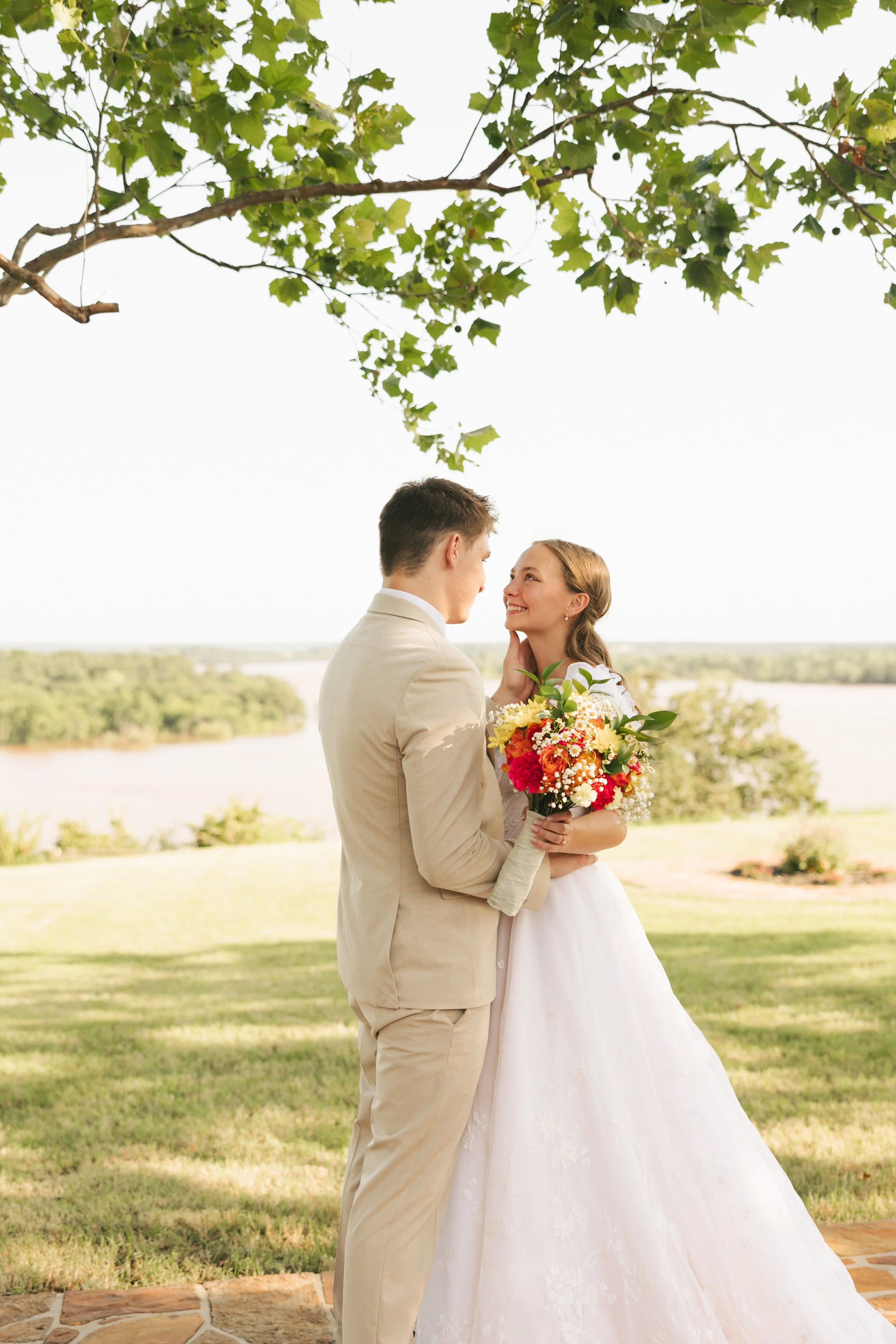 A bride and groom standing outdoors beneath a tree, gazing into each other's eyes, with a scenic lake front and green landscape in the background. The bride holds a bouquet of colorful flowers.