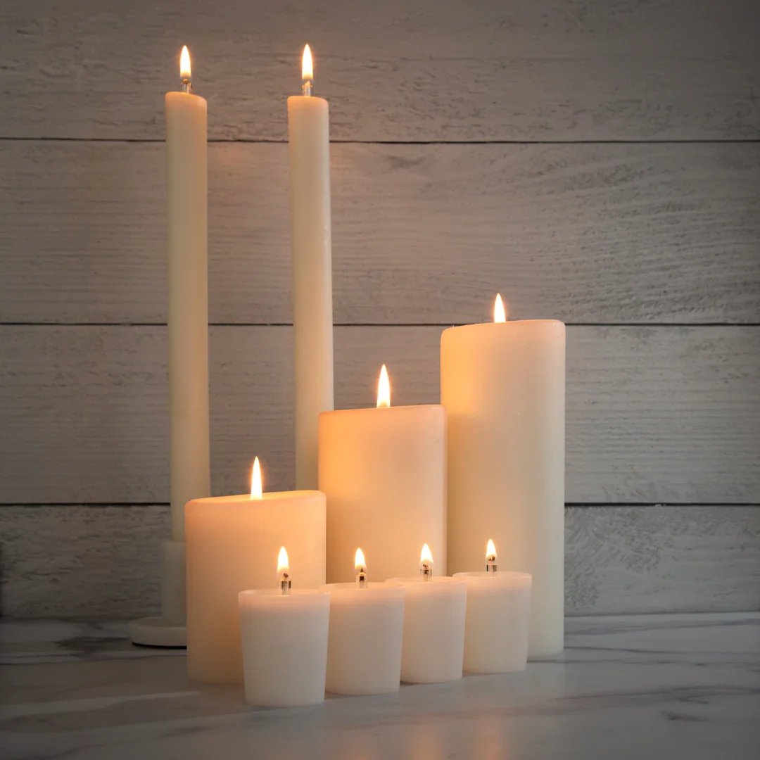 Several lit white candles of varying heights arranged outdoors against a wooden wall, creating a warm, cozy ambiance.