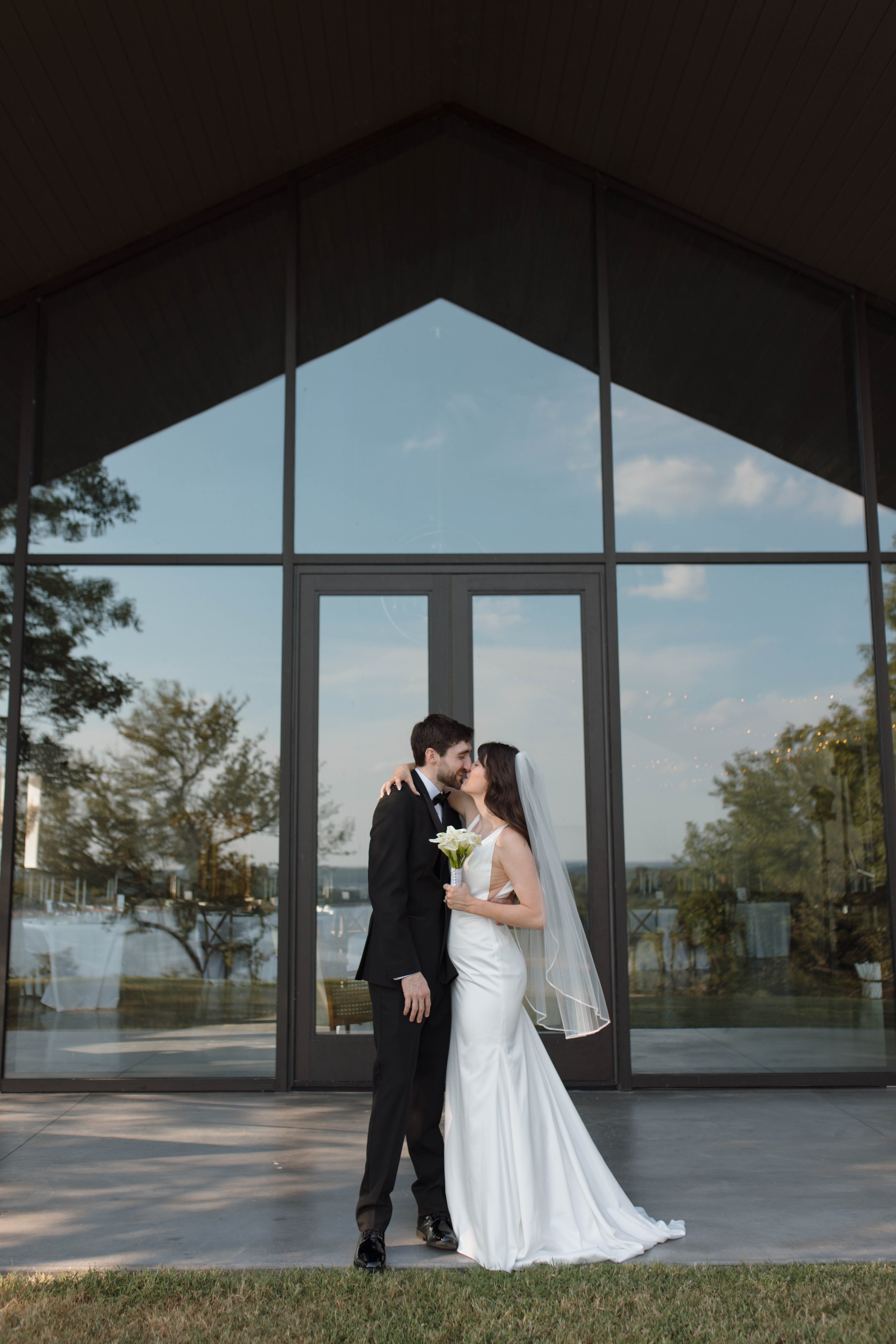 Bride and groom kissing in front of a large glass building, the bride holding a bouquet of white flowers, outside during daytime.