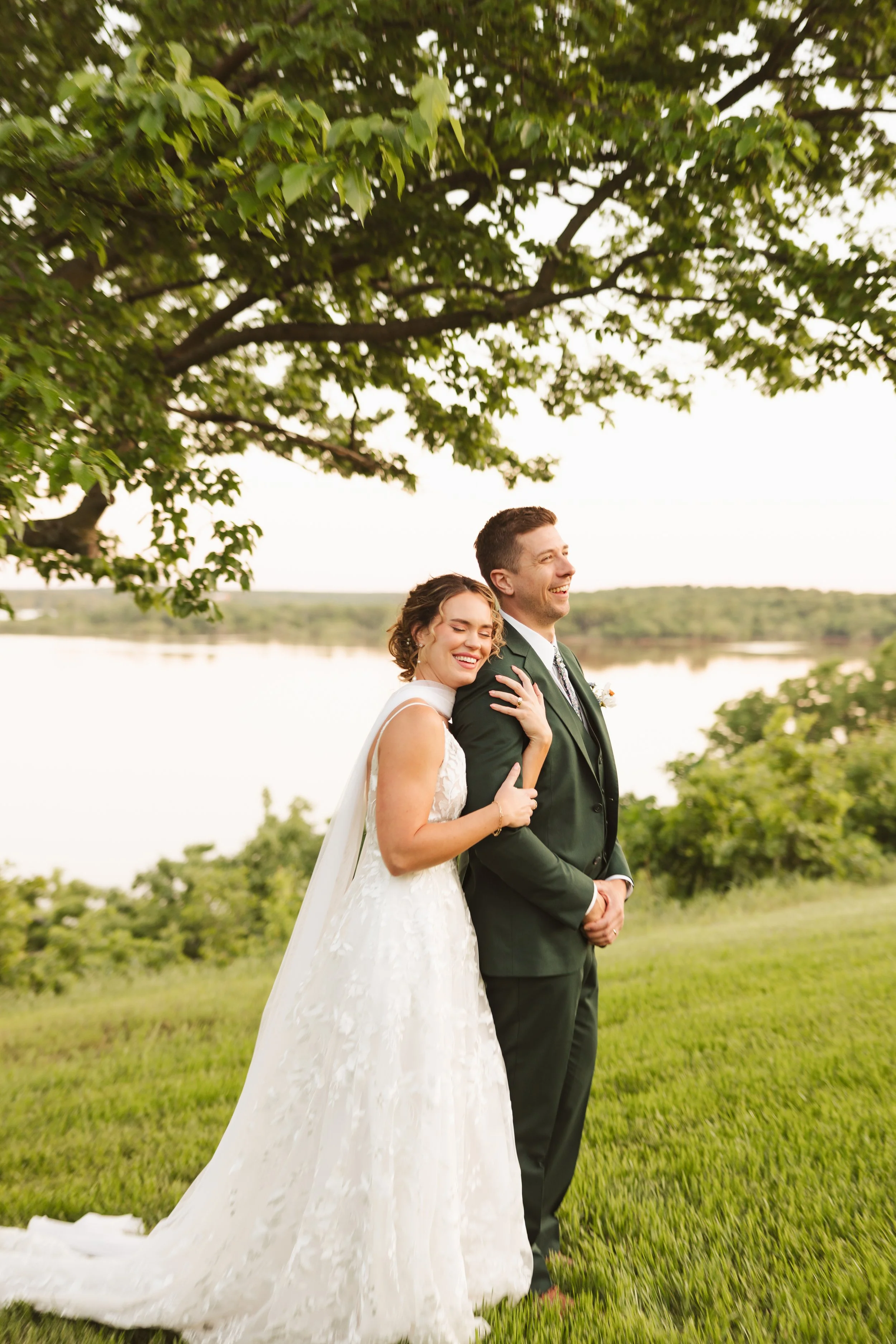 A newlywed couple in wedding attire sitting on the grass outdoors during late afternoon. The groom is pouring rosé wine into a glass held by the bride, both are smiling and enjoying the moment. The background features trees and natural sunlight.