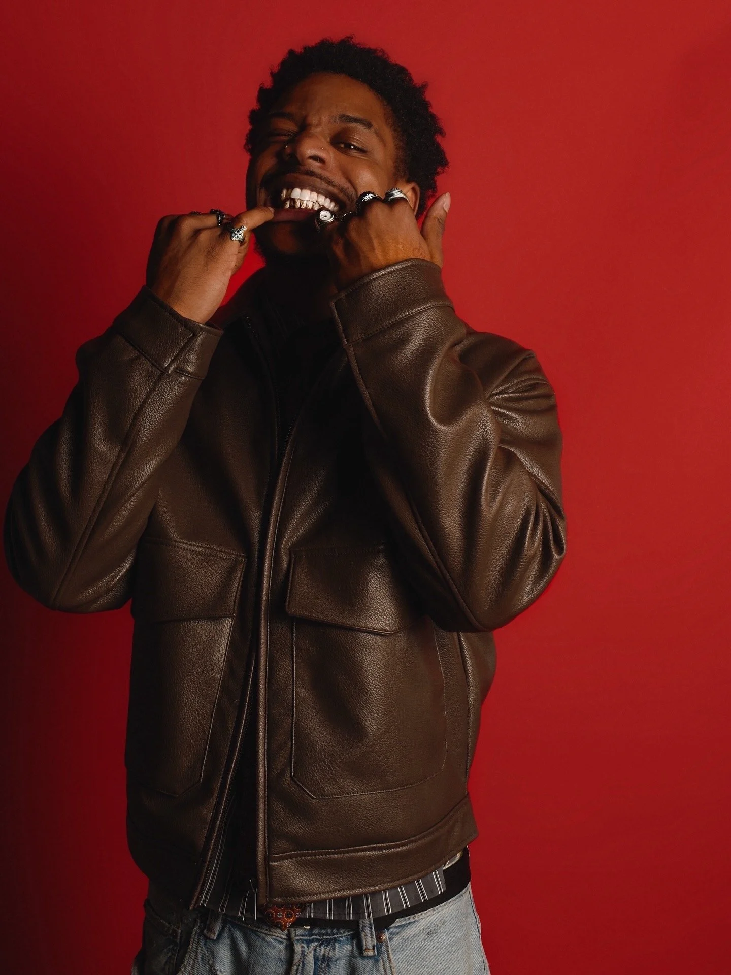A man with dark curly hair is smiling and biting his finger, wearing a brown leather jacket and jewelry, standing against a red background.