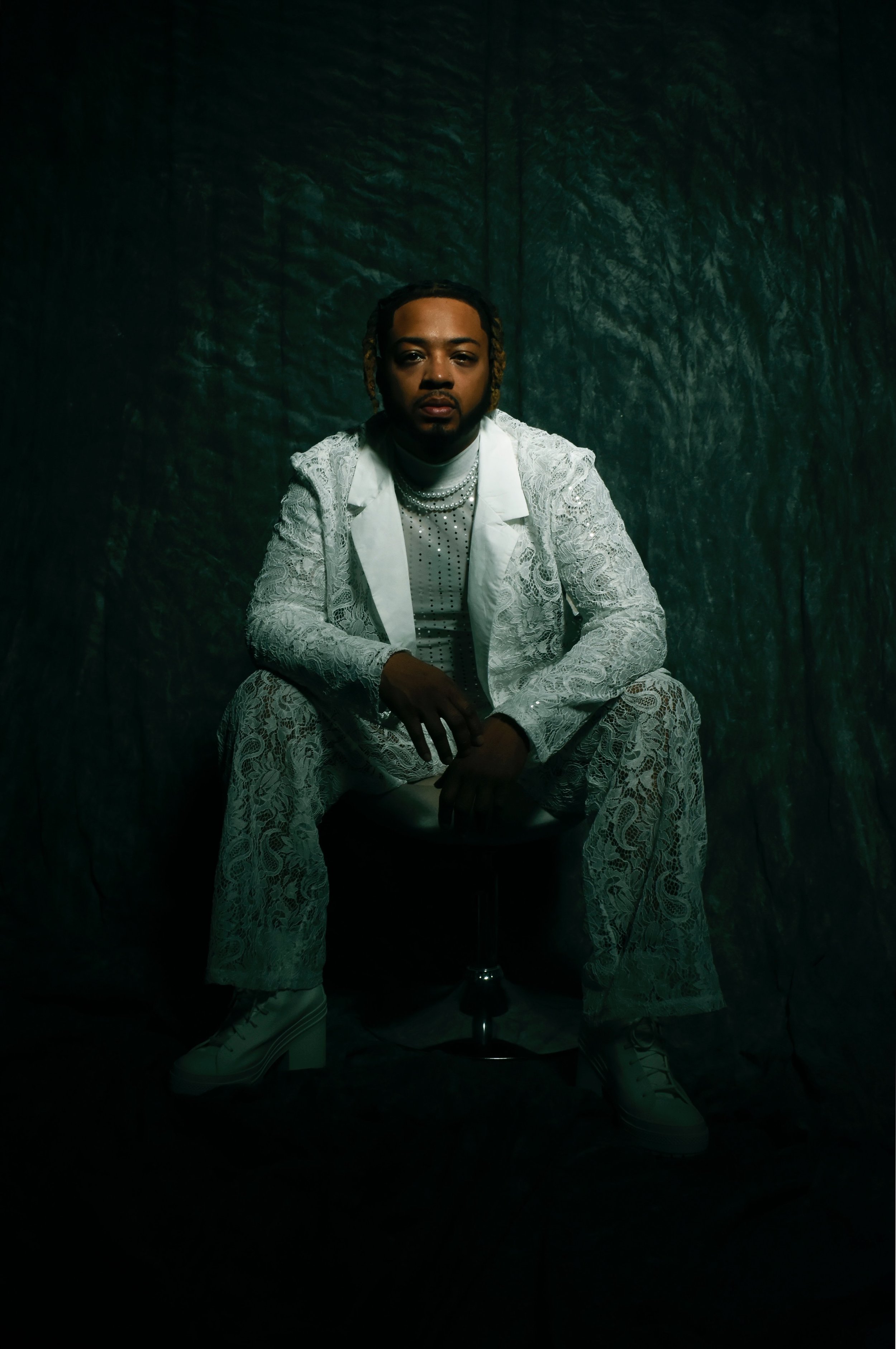 A person sitting on a stool in a dark setting, wearing a white lace suit, white shoes, and accessorized with layered necklaces, with a dark textured background.