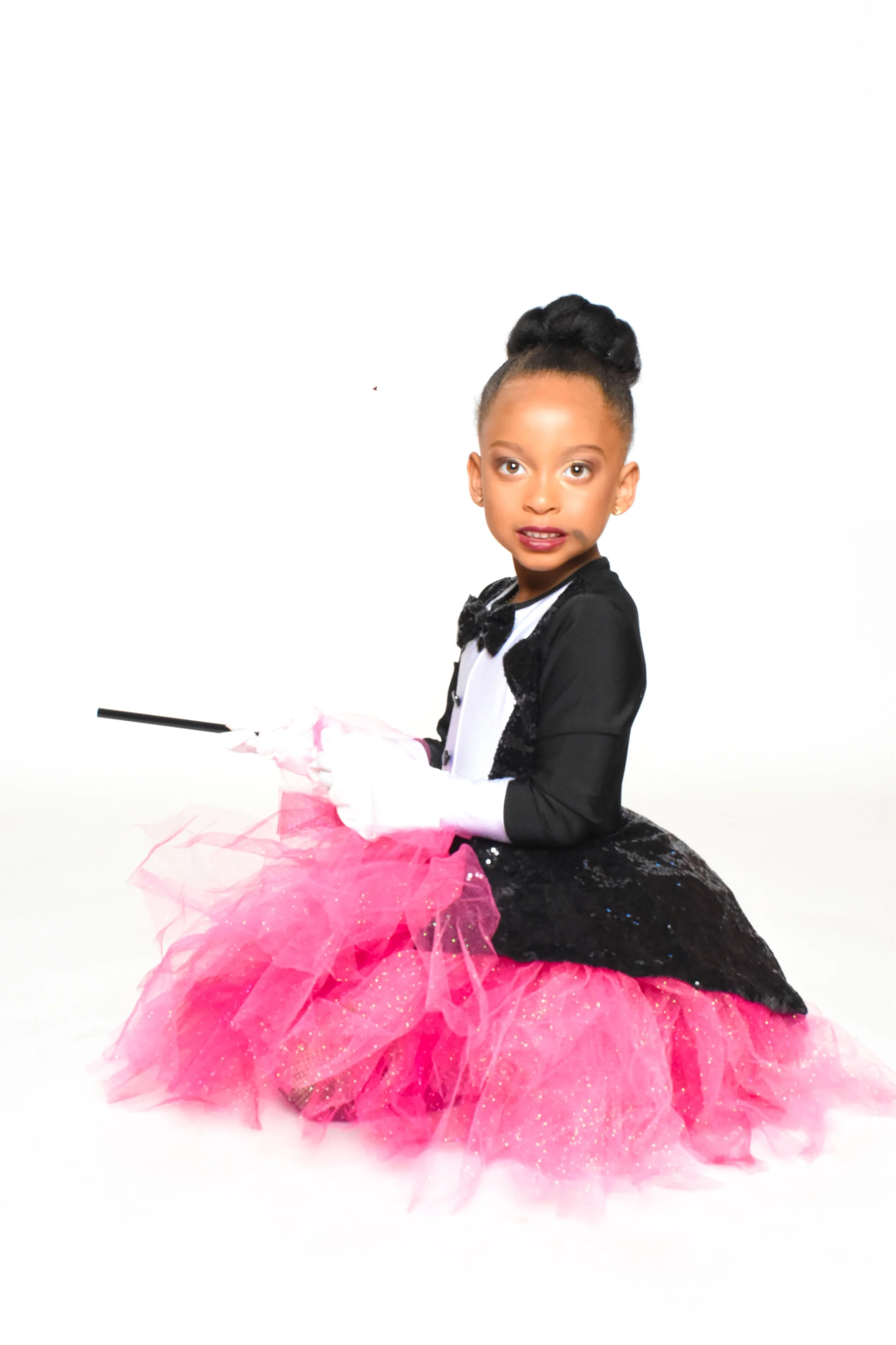 Young girl dressed in a black and pink tutu costume, sitting on the floor with a white background, holding a magic wand and wearing white gloves.
