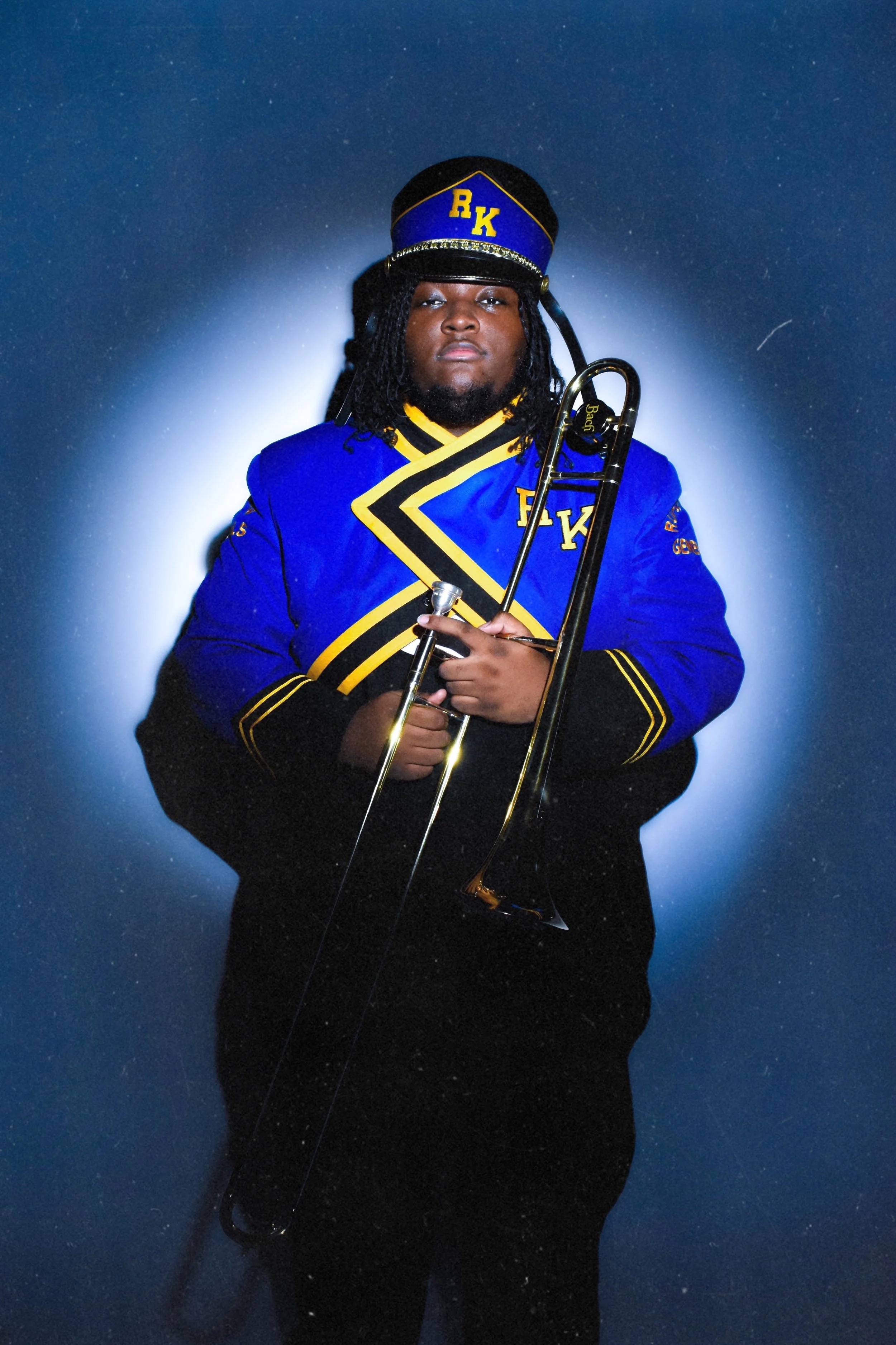 A person wearing a blue marching band uniform and cap with gold and black accents, holding a trombone, standing against a dark background with a white glow behind them.
