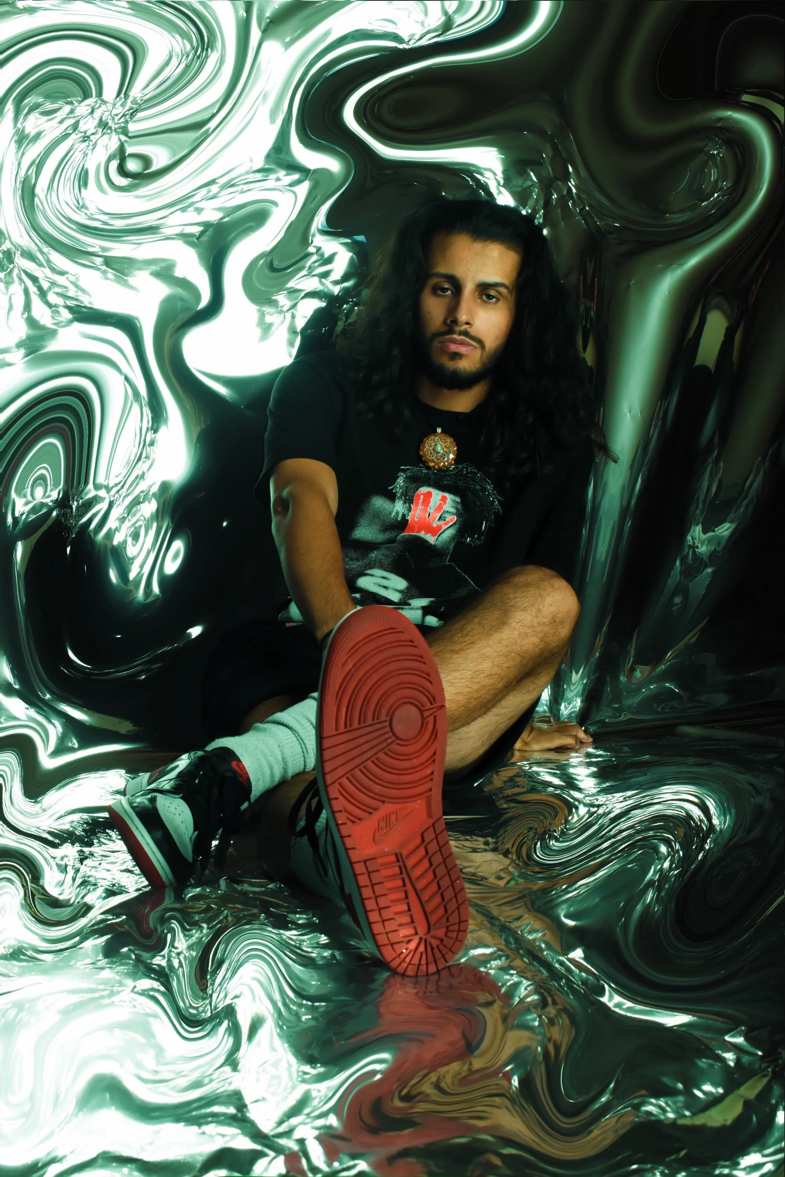 A man with long, dark hair and a beard seated on a shiny, swirling metallic surface background, wearing a black T-shirt, gray socks, and sneakers, looking at the camera with a serious expression.