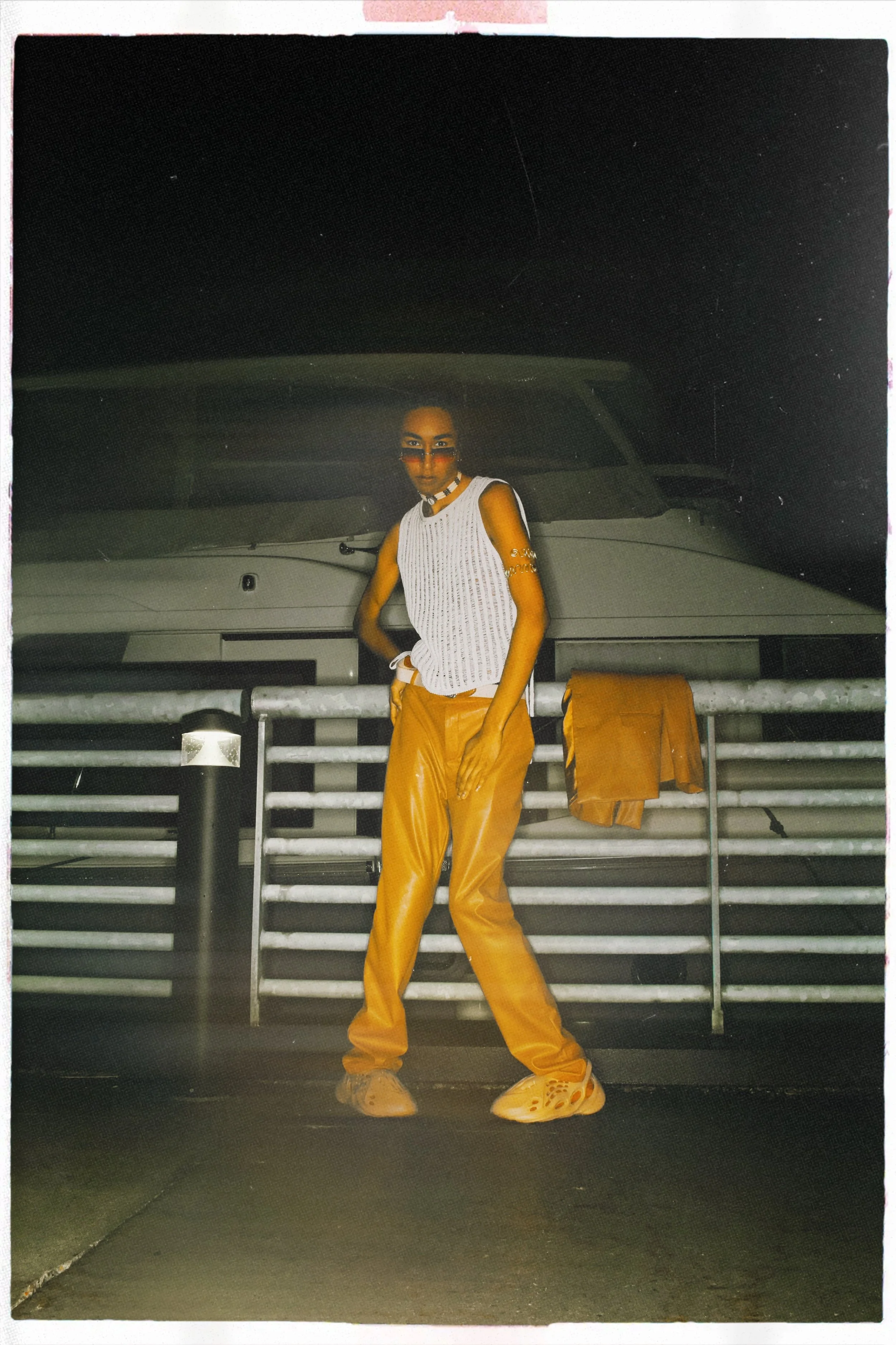 A woman in a white sleeveless top and orange pants standing in front of a boat with a metal fence, at night.
