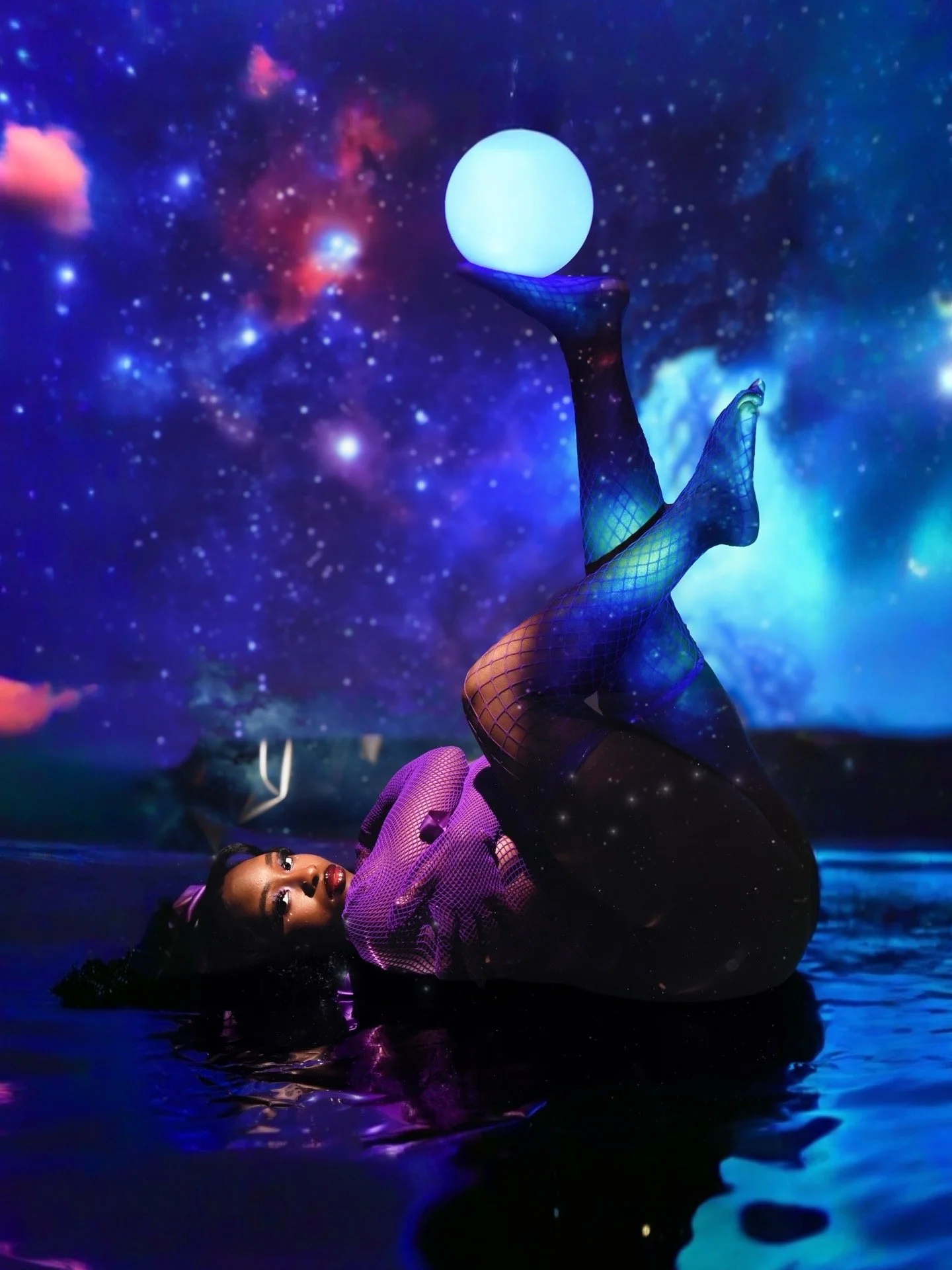 A woman lying on her side in a body of water with her legs raised, holding a glowing orb over her foot. The background features a cosmic, star-filled scene with vibrant colors.