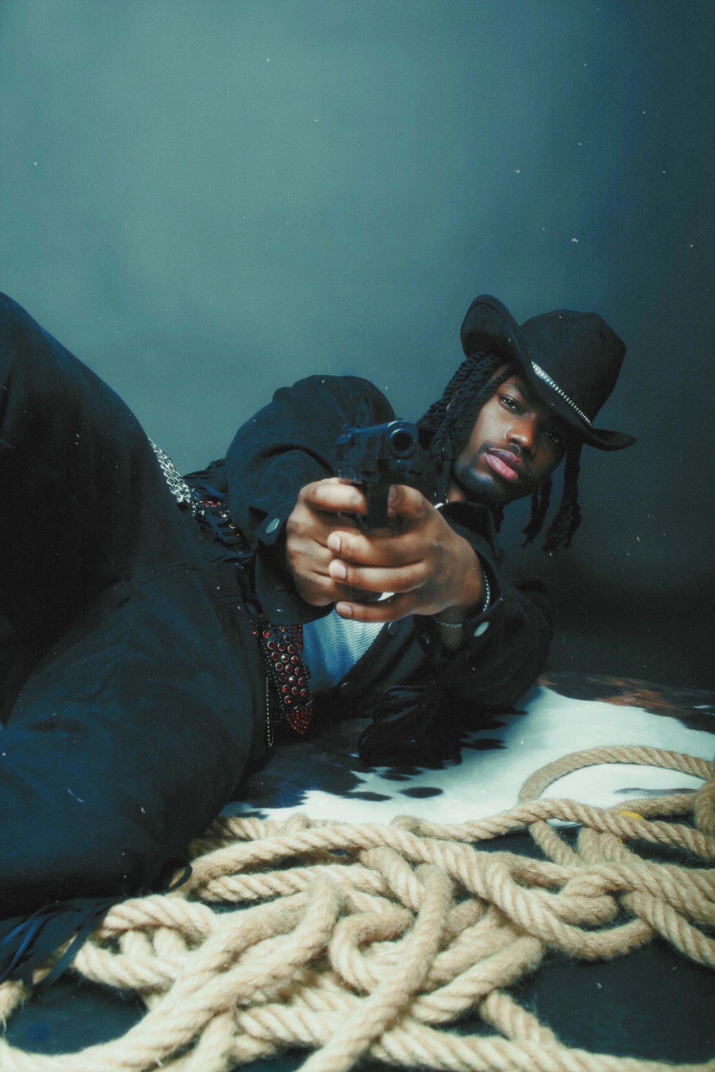 A man dressed in black lying on the floor with a thick rope in front of him, pointing a handgun at the camera, wearing a black cowboy hat and black jacket, with long dreadlocks.