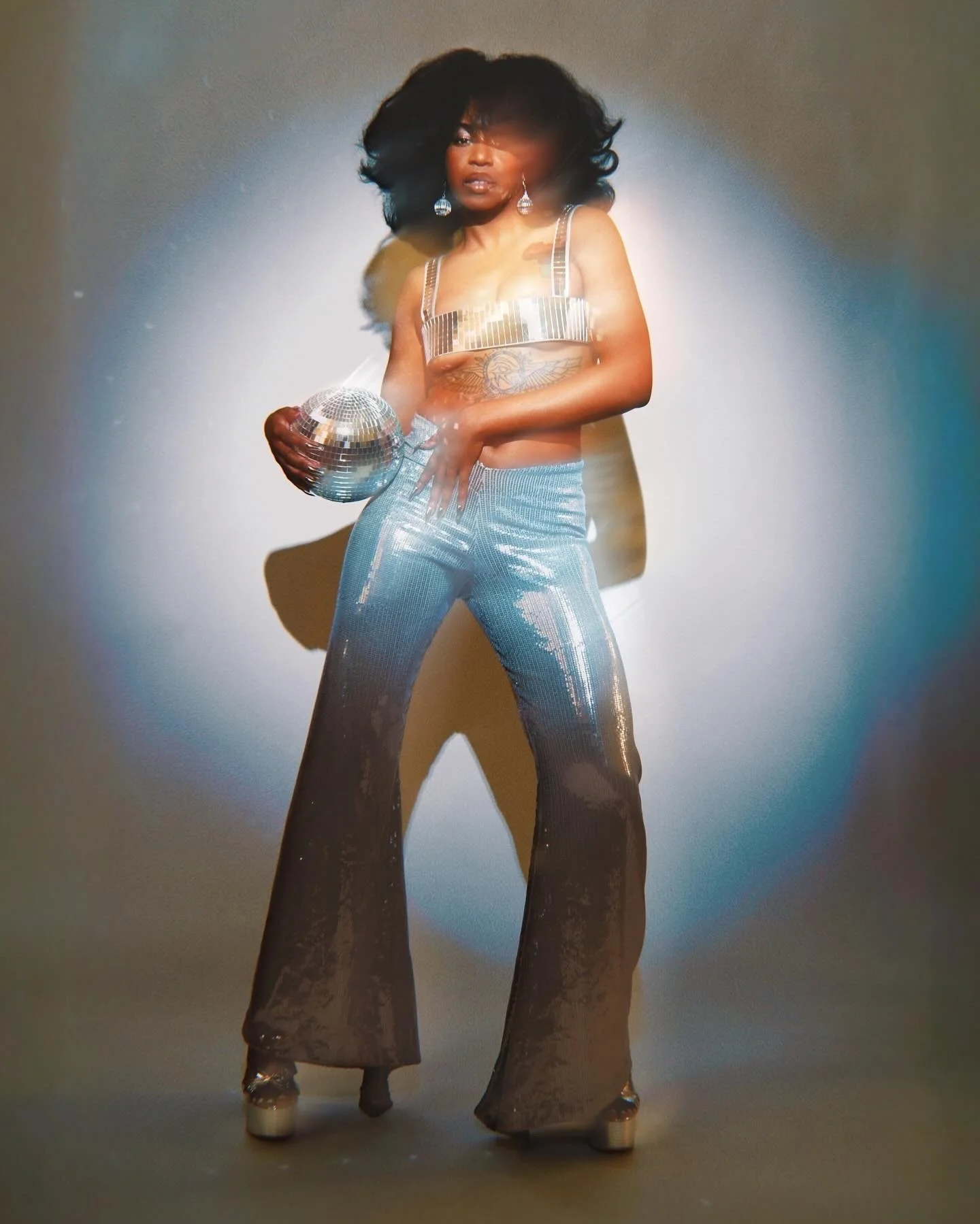 Woman in shiny silver bell-bottom pants and a silver crop top holding a mirrored disco ball, posing in front of a plain background