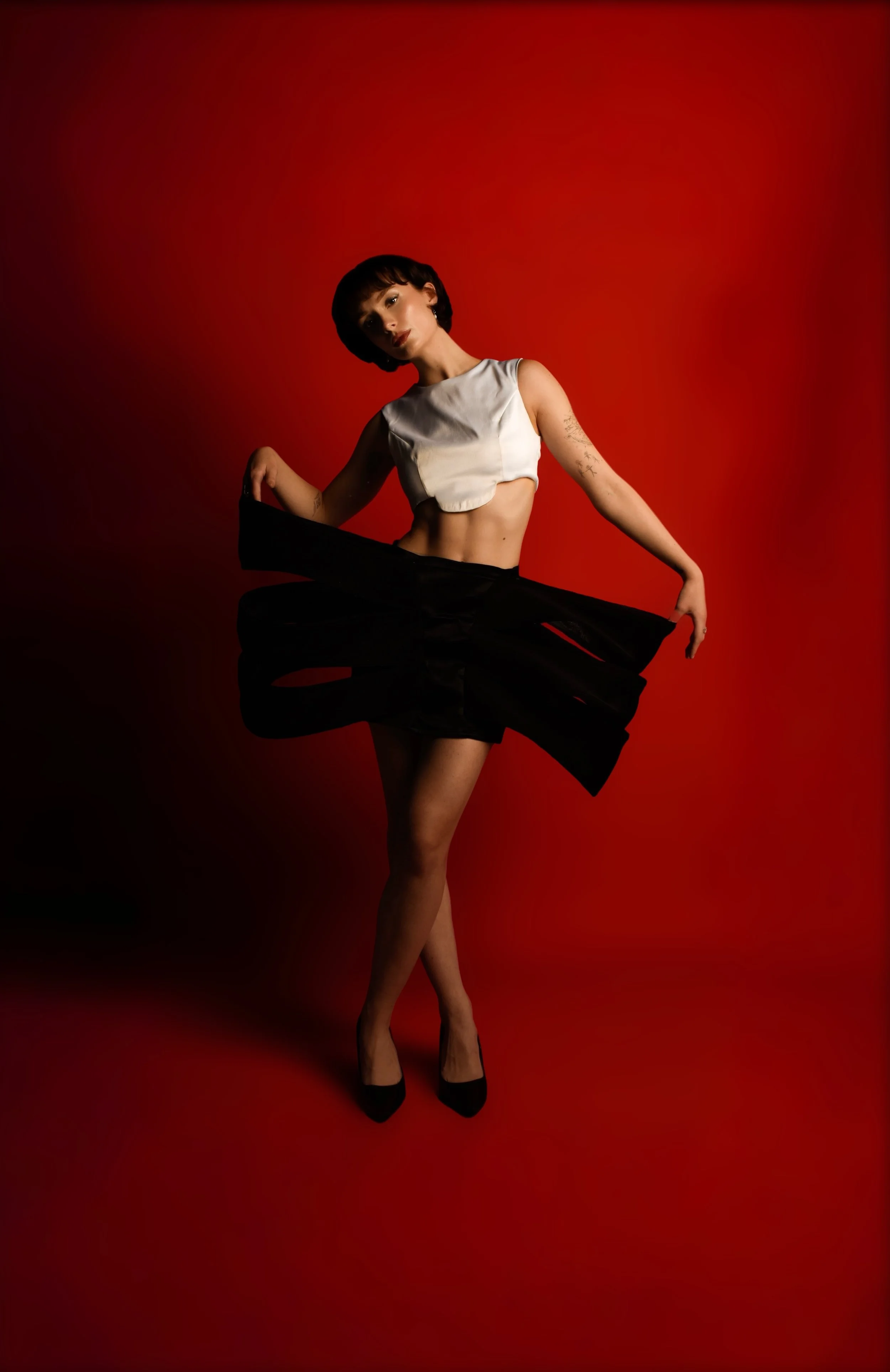 A woman with short dark hair, wearing a white crop top, black skirt, sheer black stockings, and black high heels, stands against a red background in a confident pose.