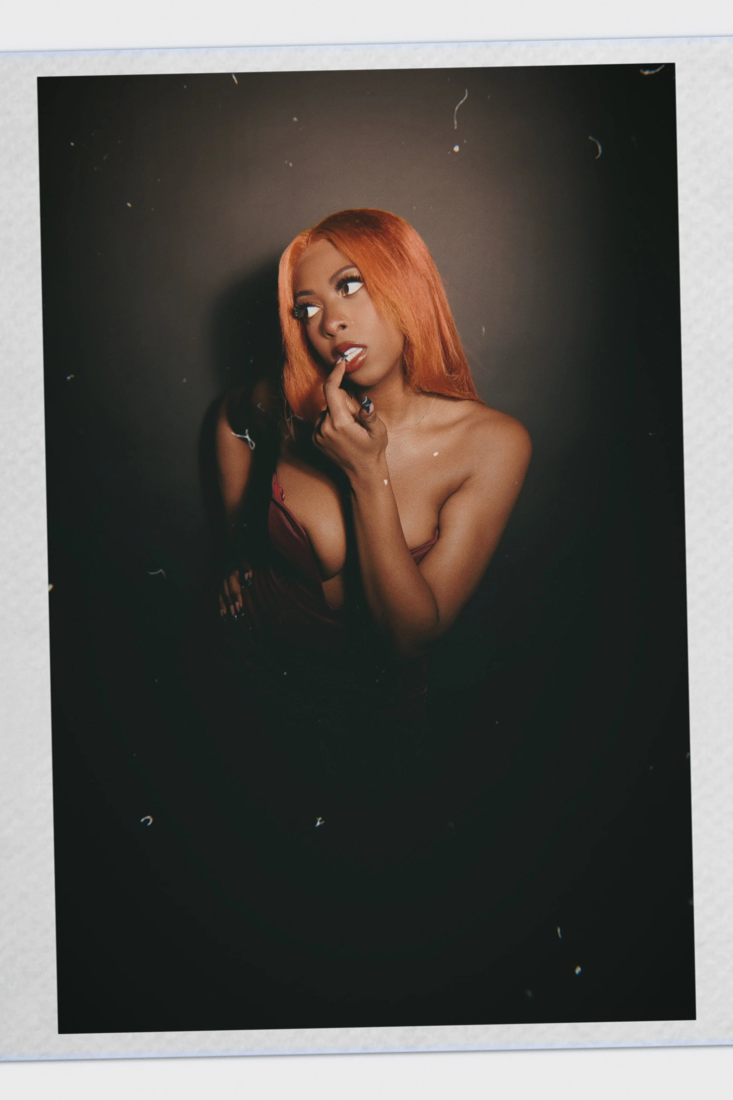 A woman with orange hair and makeup, wearing a low-cut top, posed with her finger on her lips against a dark background.